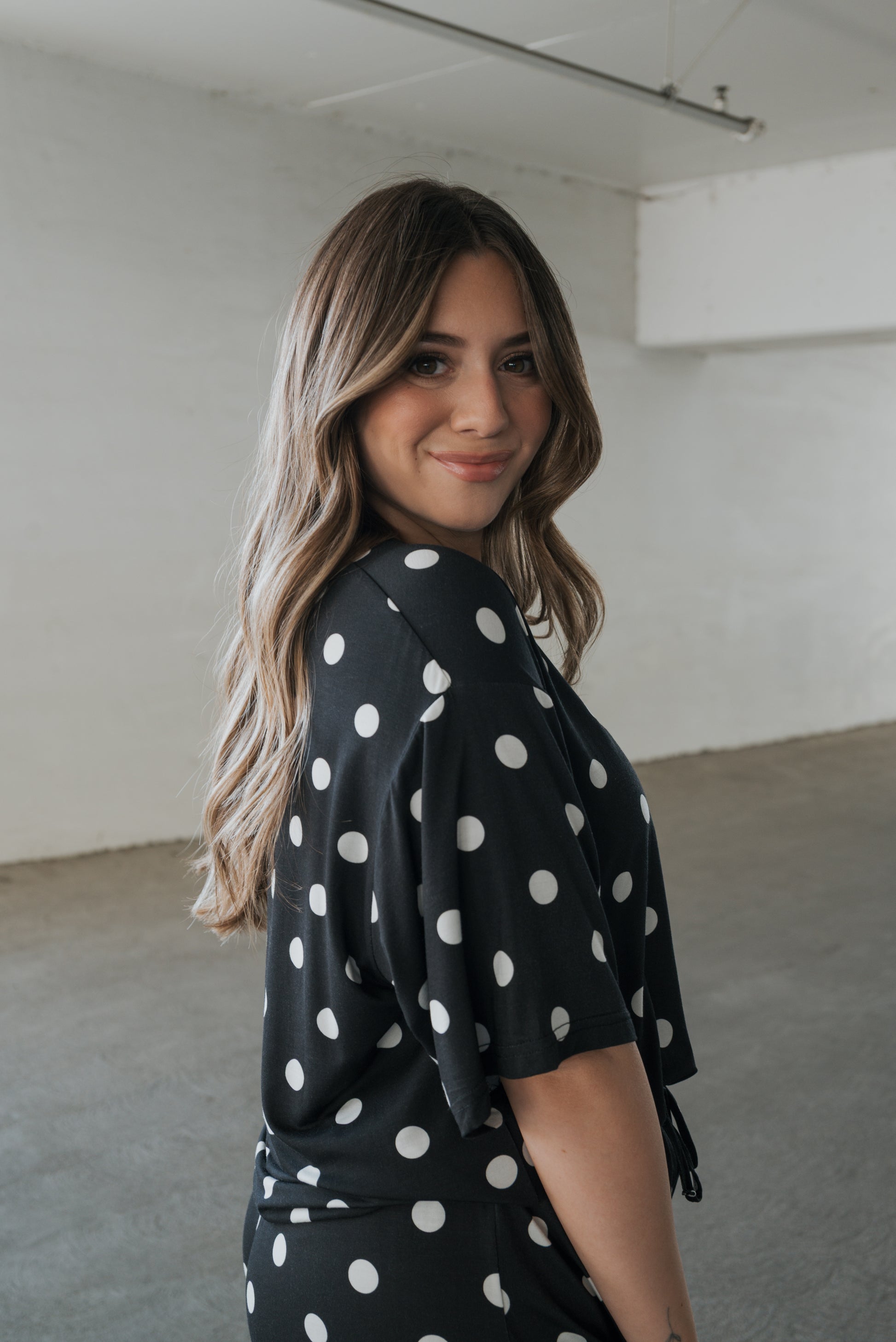A woman with long, wavy hair smiles in a minimalist, light-filled room while wearing the forever french baby Women’s Bamboo Short Set in B&W Polka Dot.