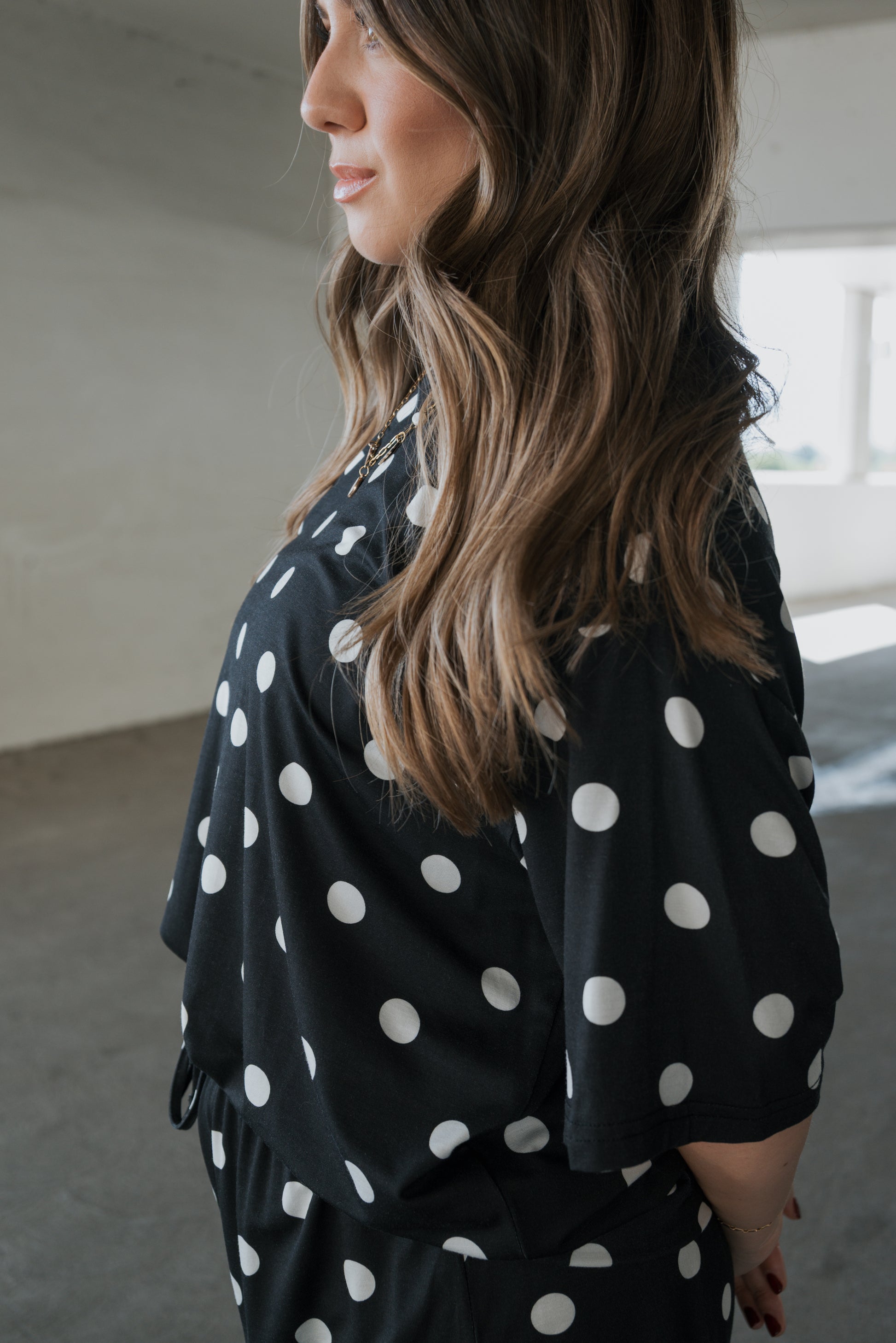 A woman with long brown hair stands in a bright room, facing sideways with her hands behind her back, wearing the forever french baby Women’s Bamboo Short Set in black and white polka dot—made from breathable bamboo fabric.