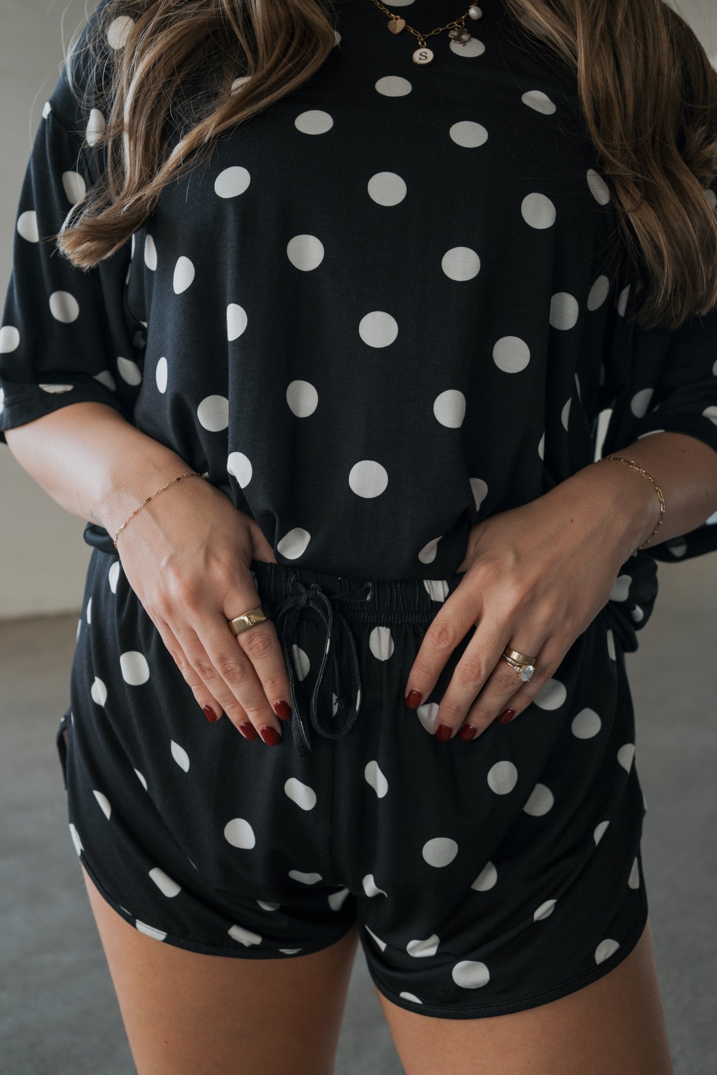A person wears the forever french baby Women’s Bamboo Short Set in B&W Polka Dot, a black breathable romper with white polka dots. Their hands rest on their waist, showing off painted nails, rings, bracelets, and a necklace. Wavy hair is partially visible.