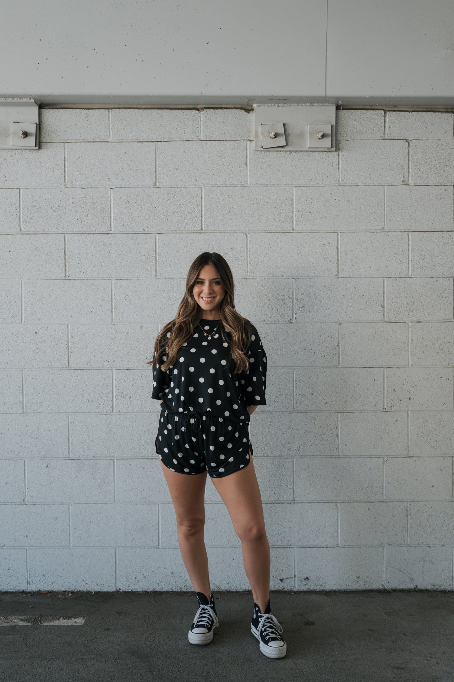A woman stands in front of a white brick wall, smiling, wearing the forever french baby Women’s Bamboo Short Set in Black & White Polka Dot along with black sneakers.