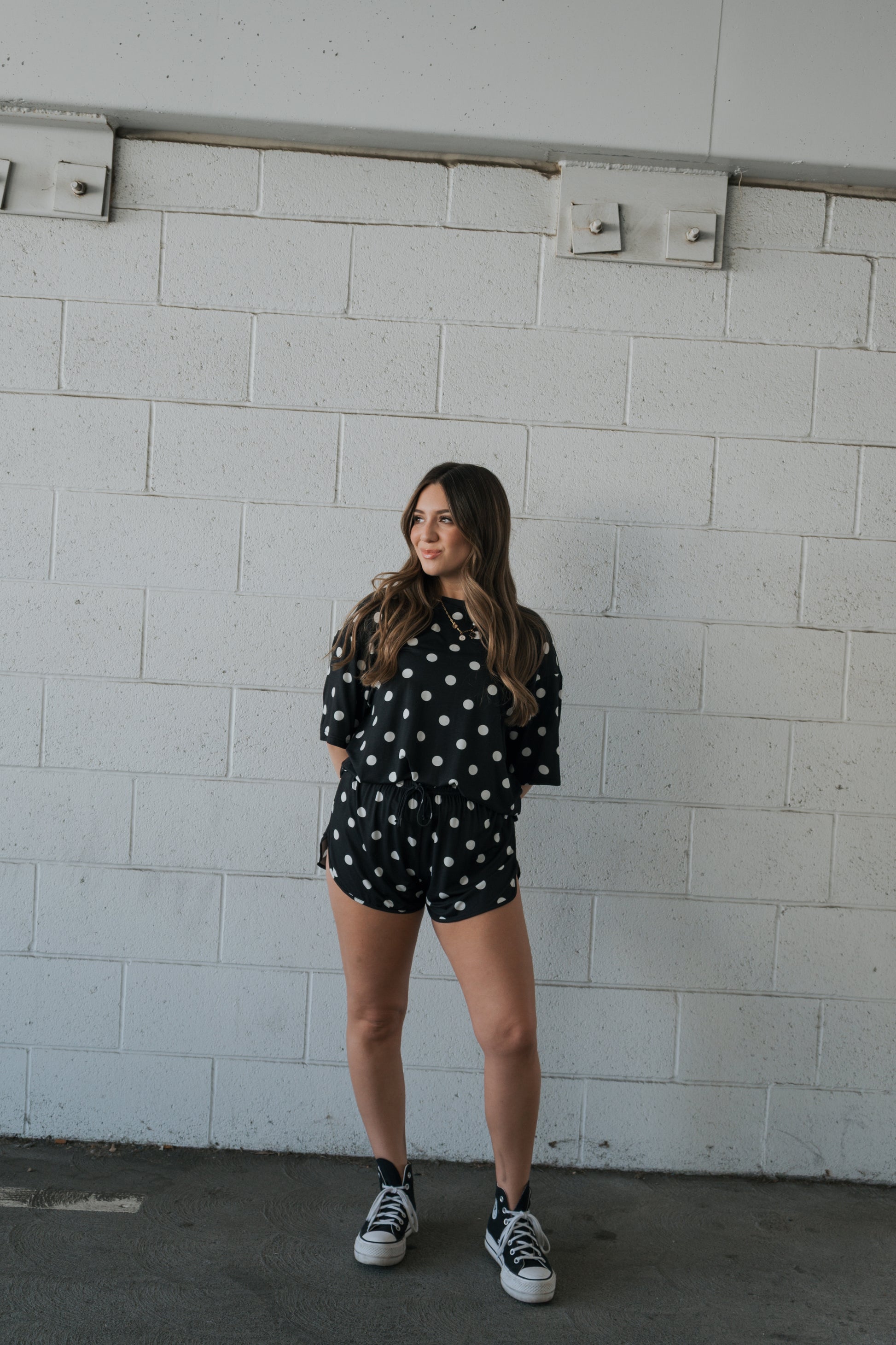 A woman stands against a white brick wall, looking to the side in effortless style. She wears the forever french baby Women’s Bamboo Short Set in Black & White Polka Dot, paired with black sneakers, her loose and wavy hair complementing the breathable bamboo ensemble.