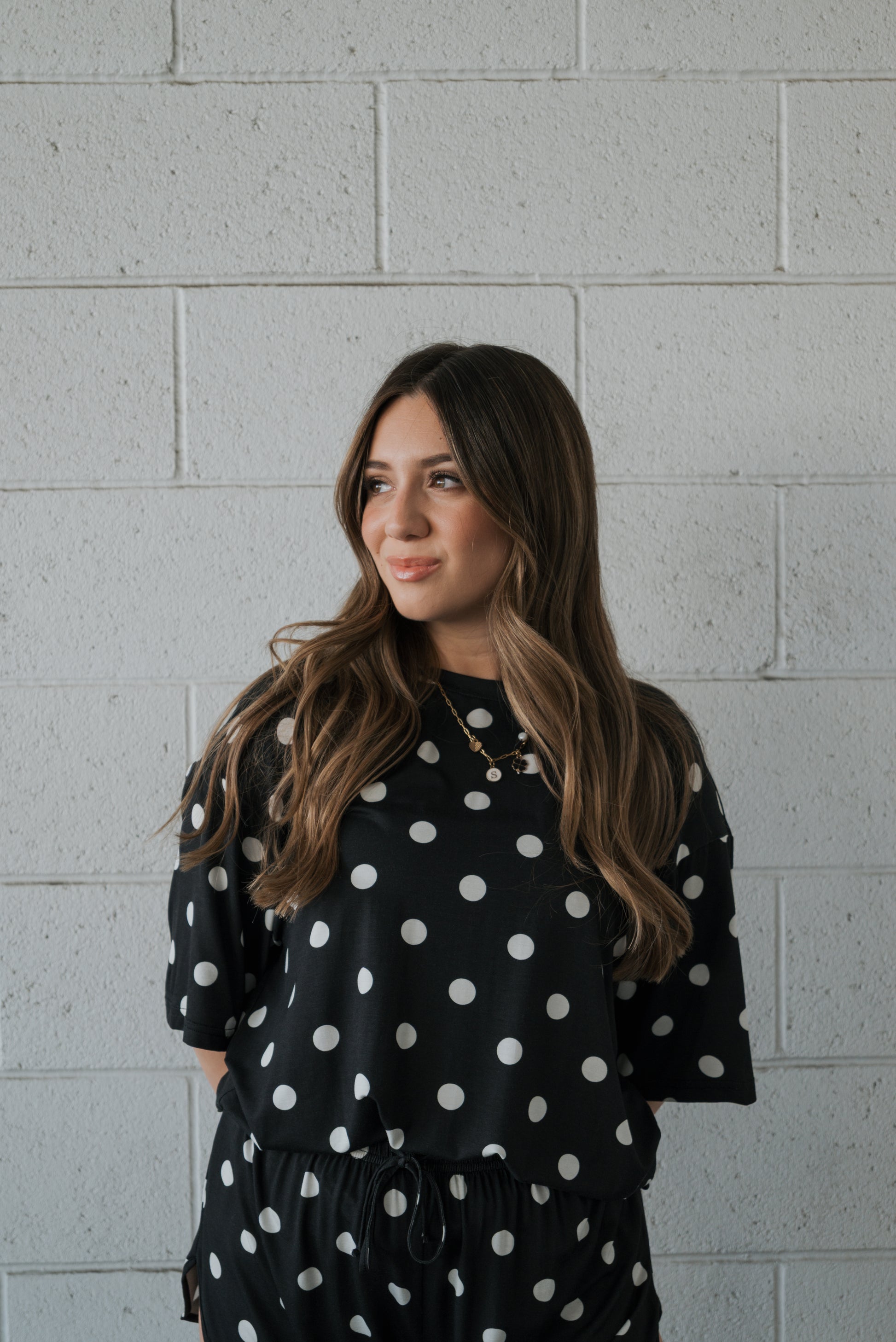 A woman with long brown hair stands against a light gray brick wall, smiling slightly to her left as she models the forever french baby Women’s Bamboo Short Set in the B&W Polka Dot print.
