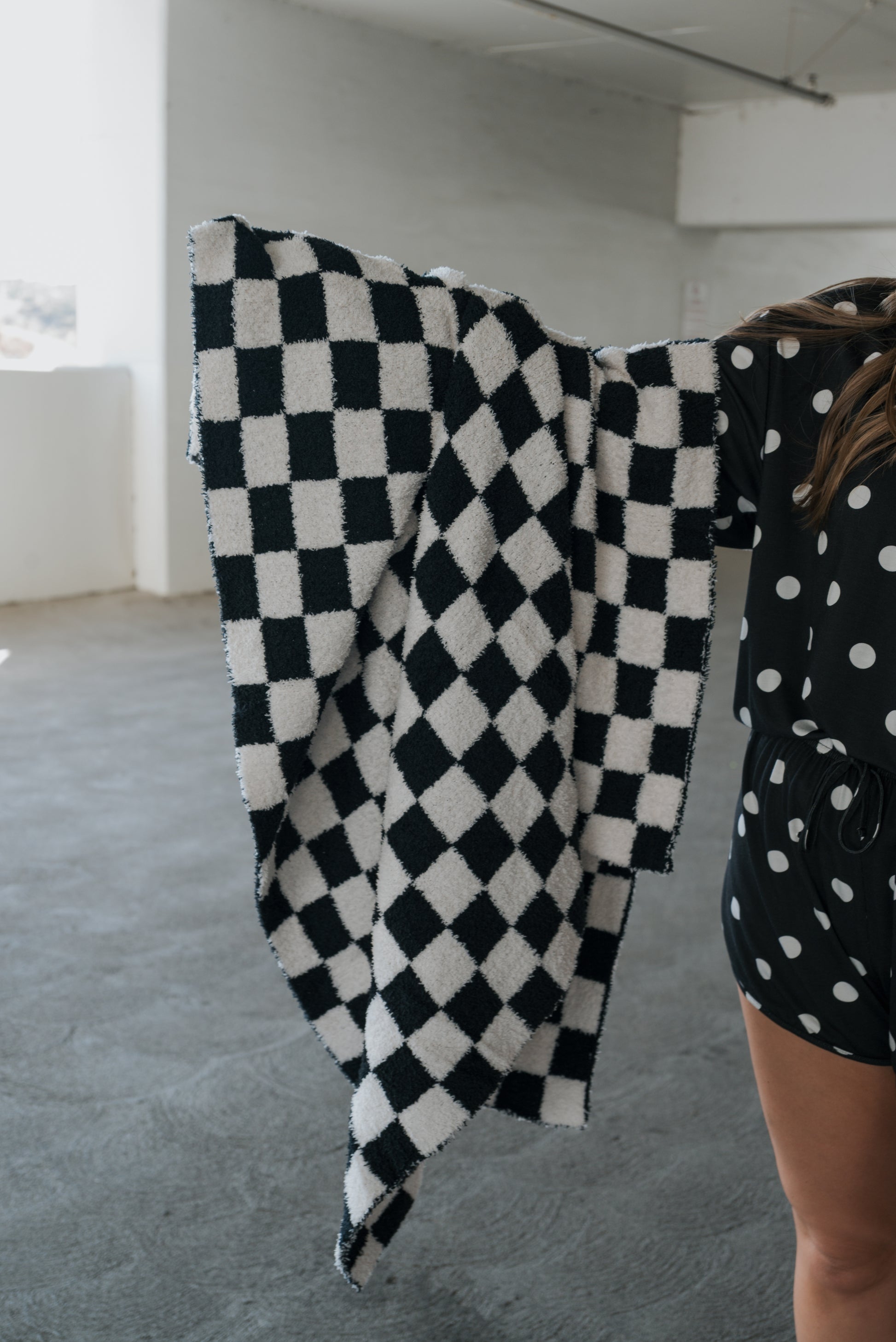 A person in a black romper with white polka dots holds the “Plush Blanket | B&W Micro Check” by forever french baby, which features an Oeko-Tex certified black and white checkered design. The scene is set in a bright, empty room with concrete floors and white walls.
