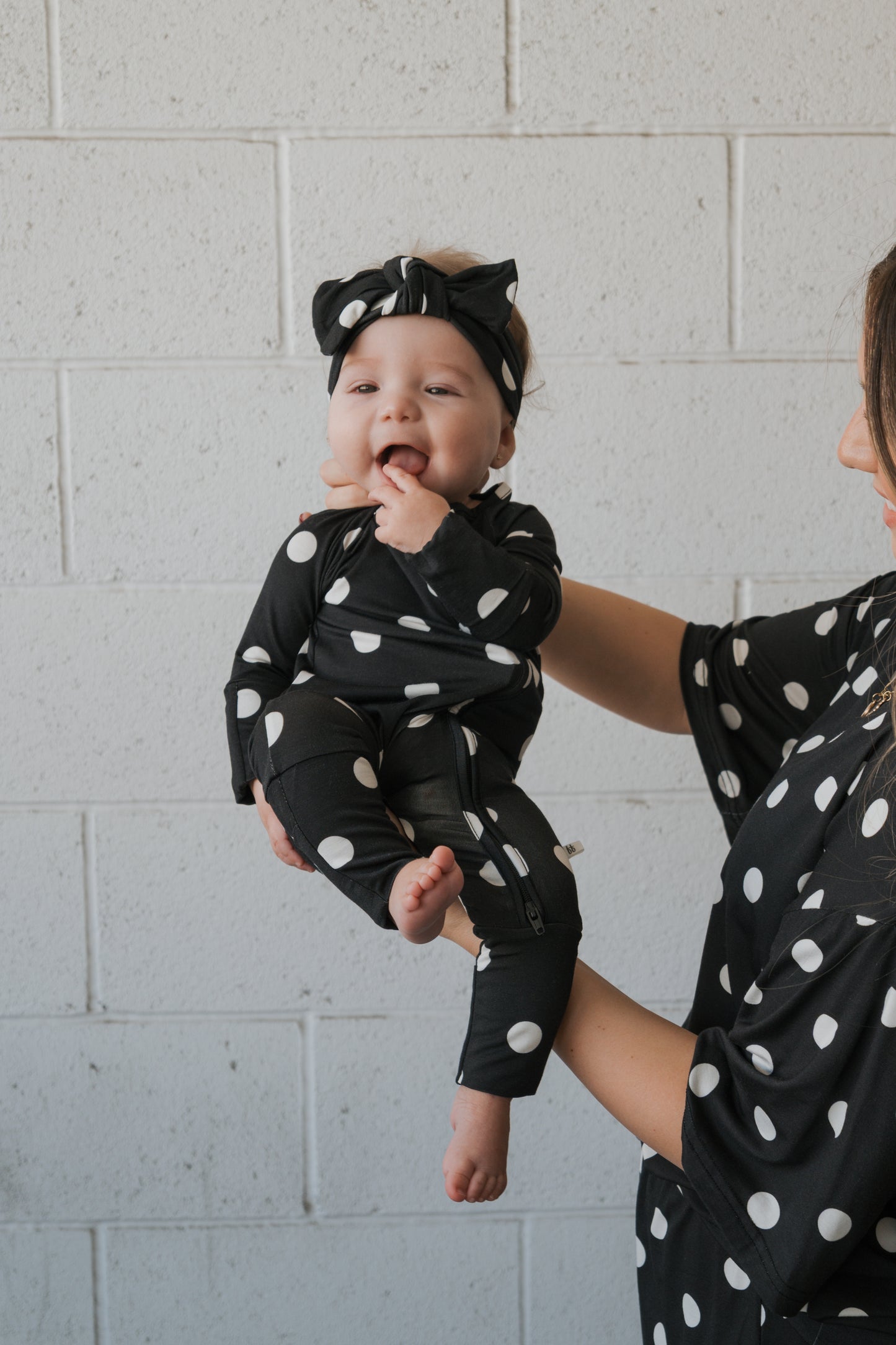 A baby dressed in forever french baby’s Bamboo Zip Pajamas in the B&W Polka Dot pattern, complete with a matching headband, is held by an adult whose arm and partial body are also clad in coordinating polka dot attire. Both wear the hypo-allergenic outfits against a white brick wall backdrop.