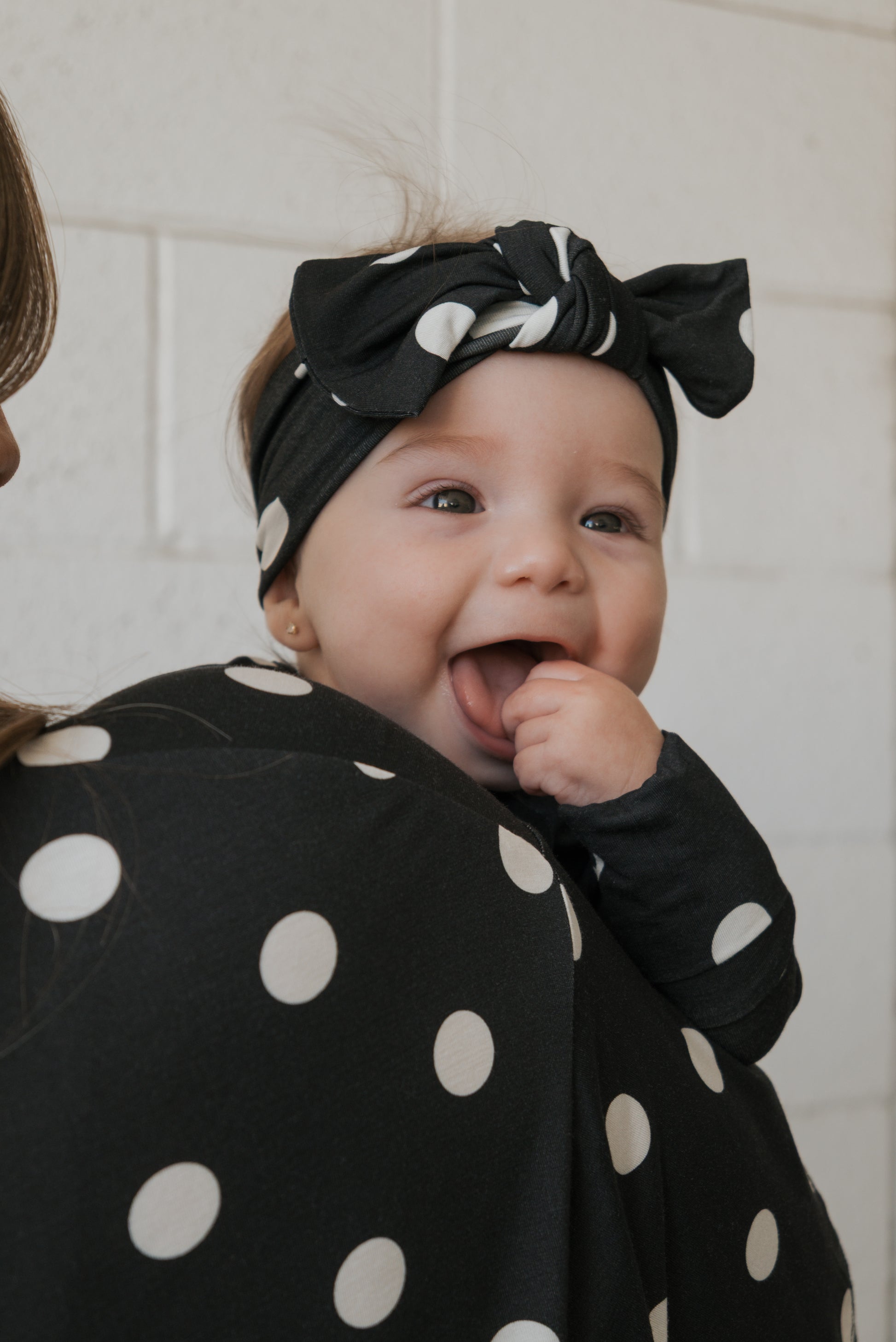 A smiling baby wearing the forever french baby Bamboo Head Wrap | B&W Polka Dot and a matching polka dot outfit sucks on their finger while being held by an adult also accessorized with a coordinating Bamboo Head Wrap | B&W Polka Dot, both showcasing the adorable forever french baby polka dot head wraps.