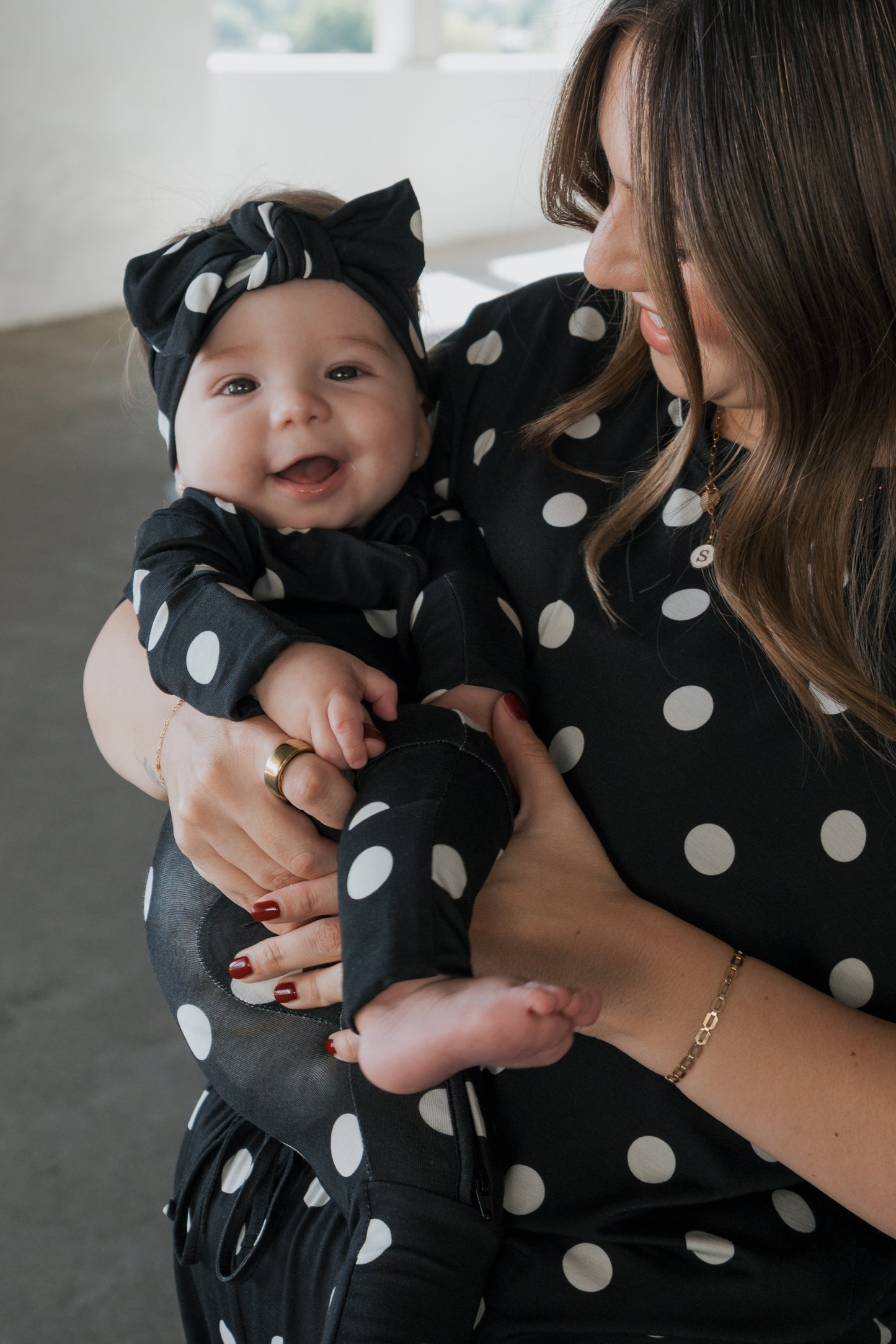 A smiling baby dressed in forever french baby’s Bamboo Zip Pajamas | B&W Polka Dot and matching headband is held by an adult with wavy brown hair, red painted nails, and a coordinating polka dot top. Both are indoors.