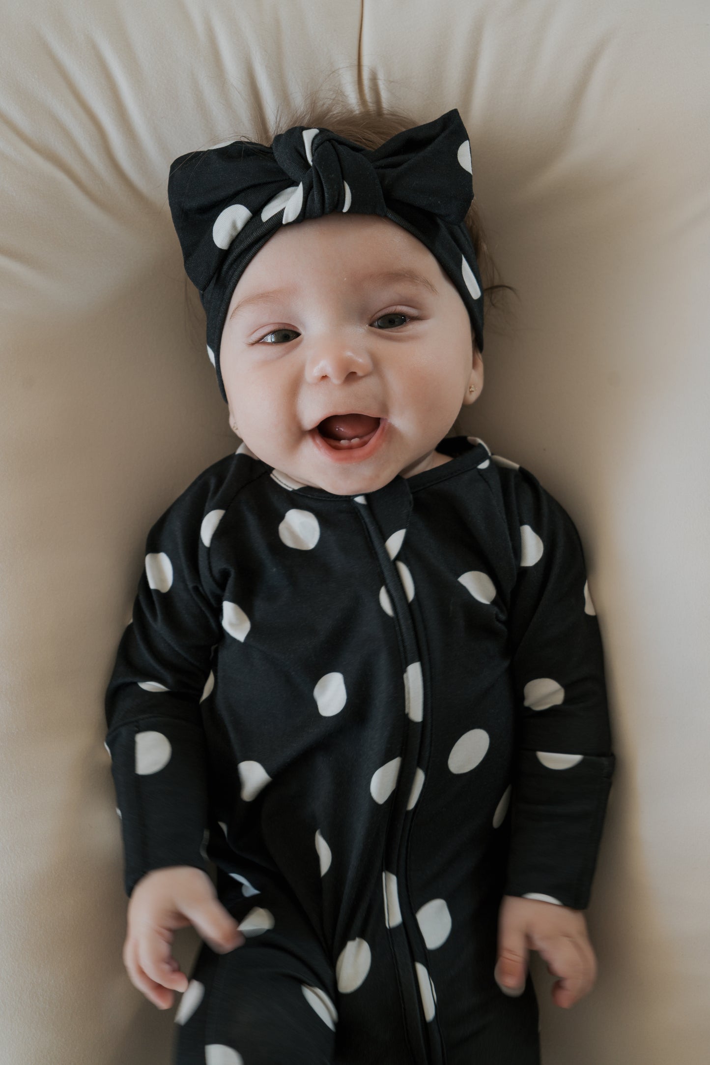 A smiling baby wearing forever french baby’s Bamboo Zip Pajamas in B&W Polka Dot, featuring a black outfit with large white polka dots and a black headband, lies on a beige cushion in hypo-allergenic baby clothing.