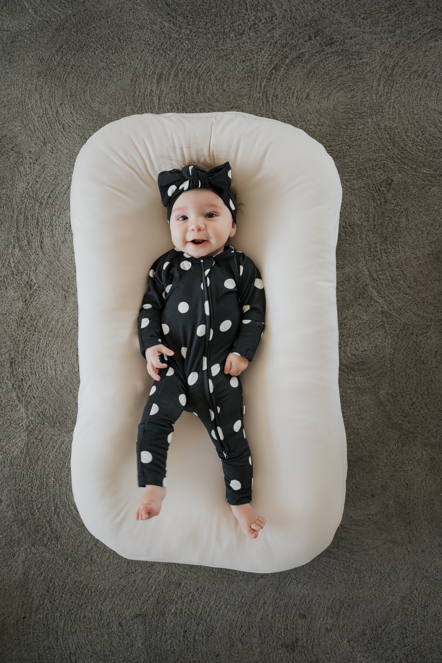 A smiling baby dressed in forever french baby’s Bamboo Zip Pajamas in B&W Polka Dot, complete with a matching headband, lies on a soft white cushion against a textured gray background.