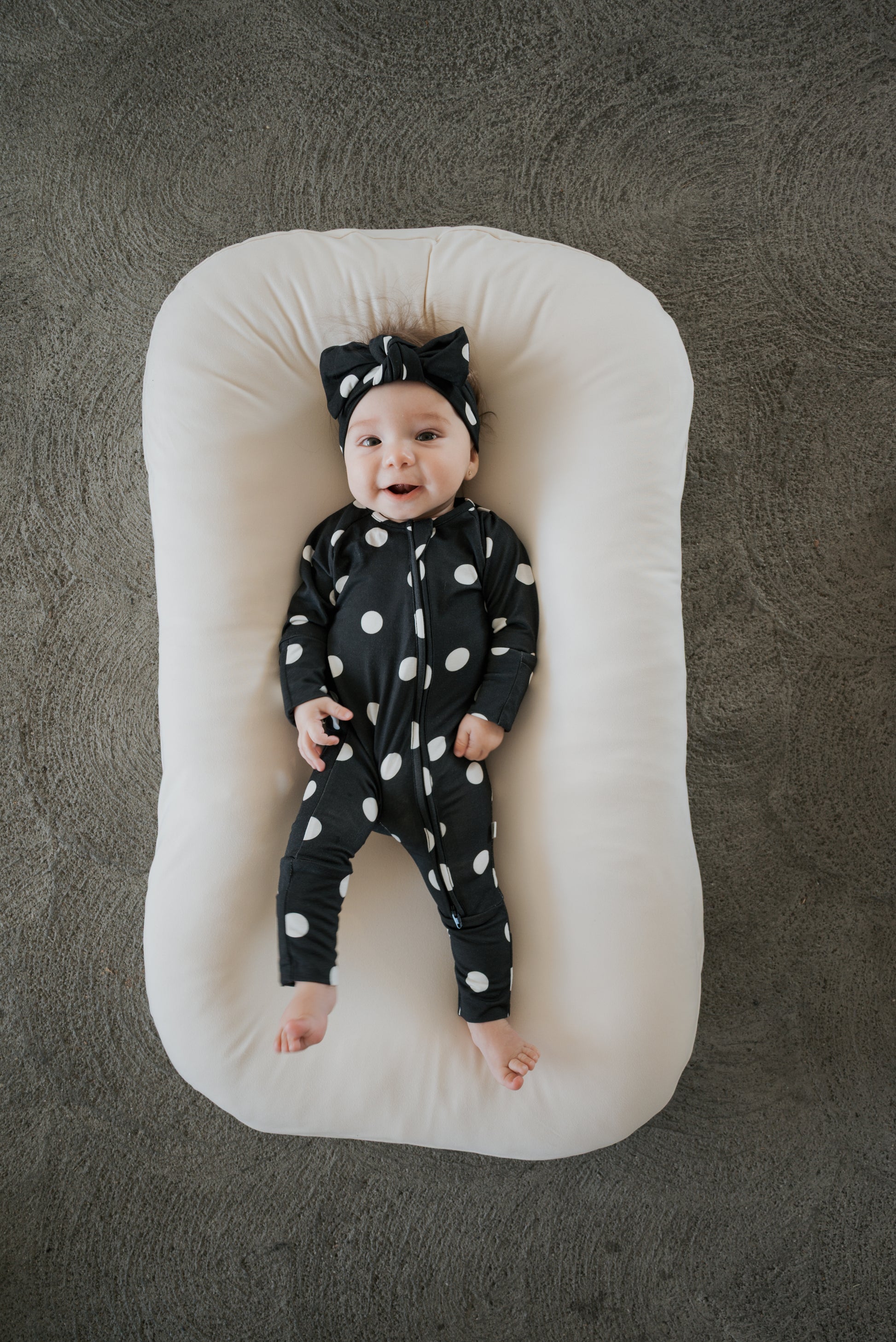 A smiling baby dressed in forever french baby’s Bamboo Zip Pajamas in B&W Polka Dot, complete with a matching headband, lies on a soft white cushion against a textured gray background.