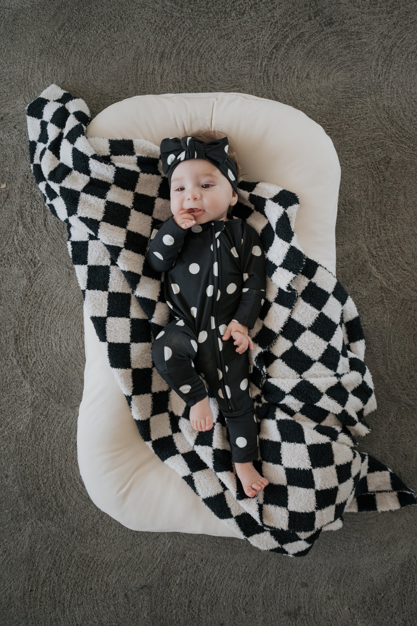 A baby in a black and white polka dot outfit and headband lies on the forever french baby Plush Blanket | B&W Micro Check, an Oeko-Tex certified microfiber blanket, which is spread over a white cushion atop a gray carpeted floor.