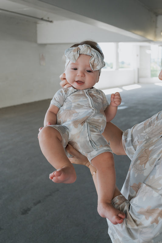 A baby wearing the forever french baby Shortie Bamboo Zip in Woodland, complete with a matching headband, is held up by an adult dressed in a similarly patterned shirt. Both display their cozy, hypoallergenic sleepwear in a bright indoor space.