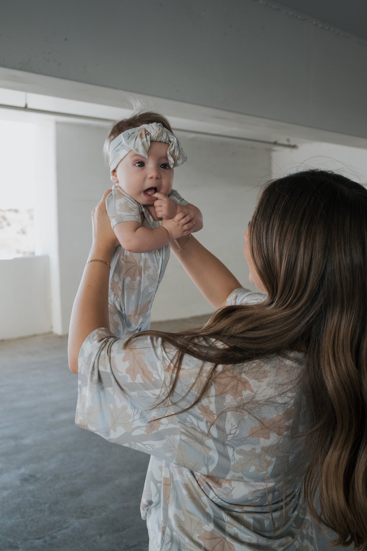 A woman with long brown hair holds up a smiling baby dressed in the forever french baby Shortie Bamboo Zip | Woodland, a floral romper and headband. Indoors, both wear matching hypoallergenic sleepwear that keeps them cozy and comfortable.