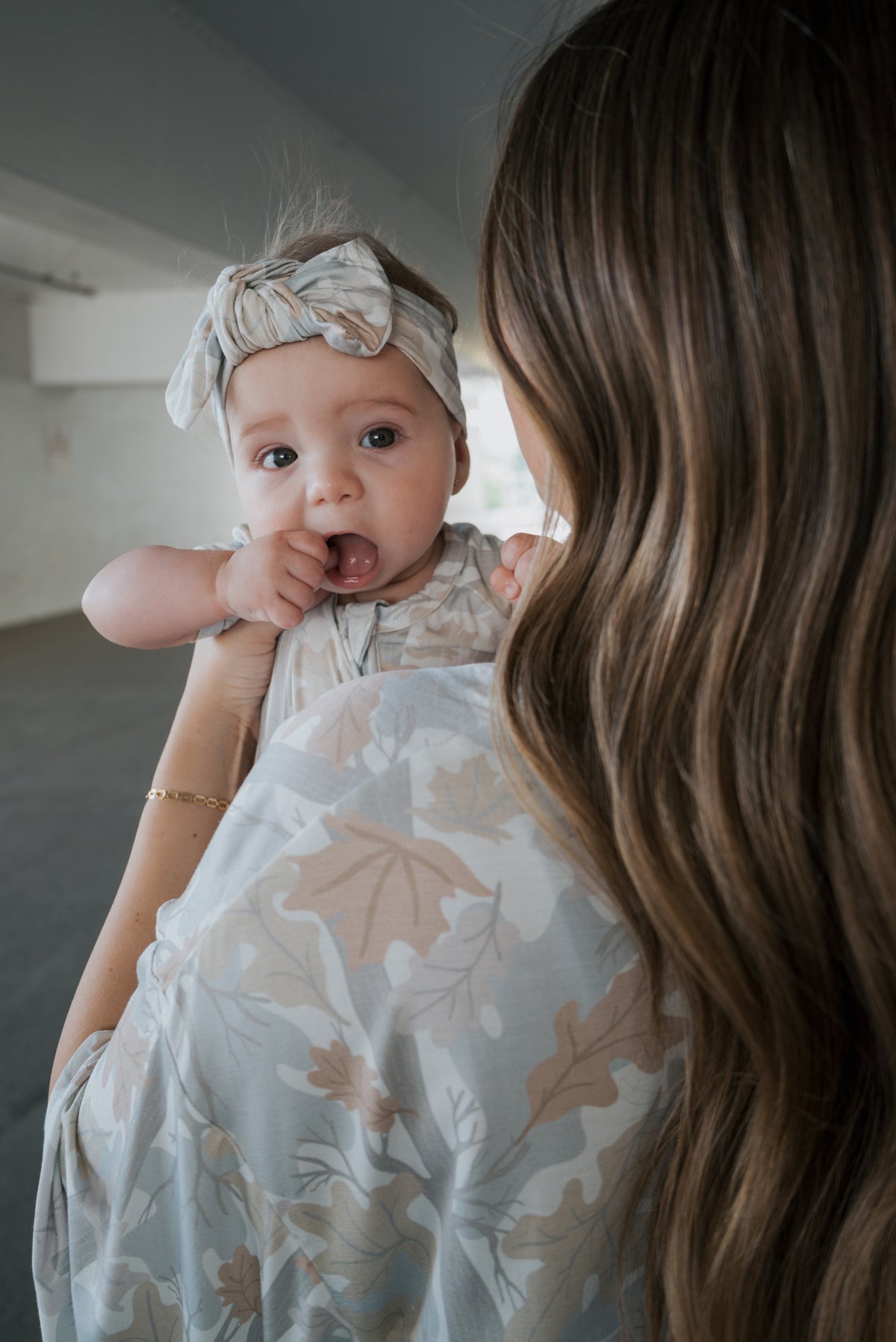 A woman with long wavy hair holds a baby wearing the Bamboo Head Wrap | Woodland by forever french baby and a patterned outfit. The baby, dressed in soft neutral tones, looks at the camera with a hand in their mouth while both are styled in gentle, earthy hues.