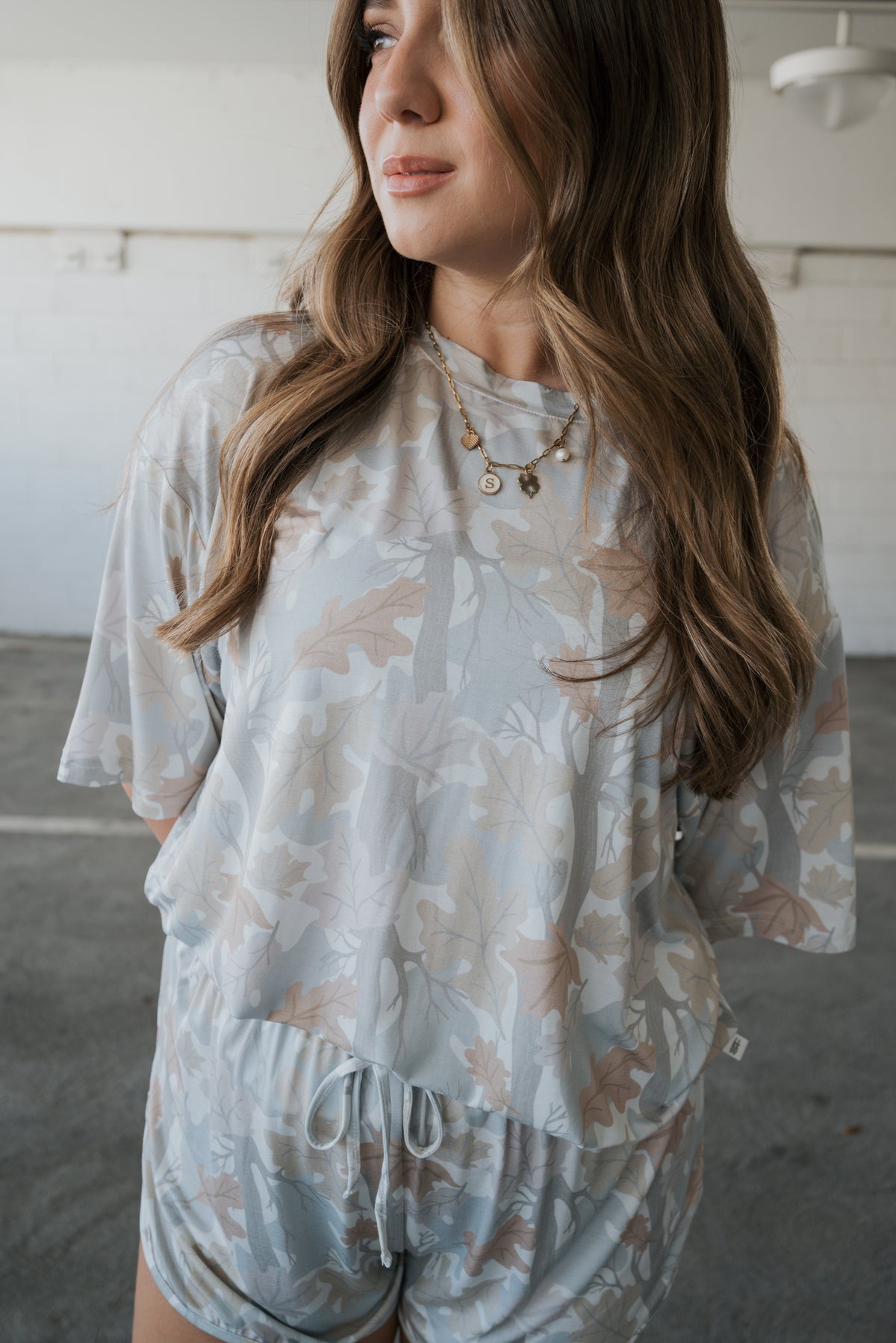 A woman with long, wavy hair stands indoors looking to the side, wearing the forever french baby Women’s Bamboo Short Set | Woodland—a matching light-colored, leaf-patterned top and shorts—paired with layered necklaces.