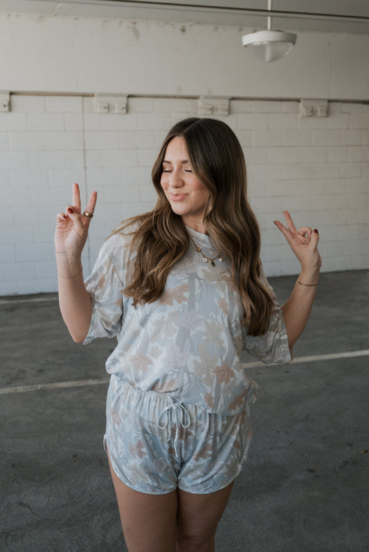 A woman with long brown hair smiles with her eyes closed and makes peace signs with both hands while wearing the forever french baby Women’s Bamboo Short Set | Woodland—a matching floral short-sleeve top and shorts crafted from ultra-soft, hypoallergenic bamboo. She stands in a light gray indoor space.