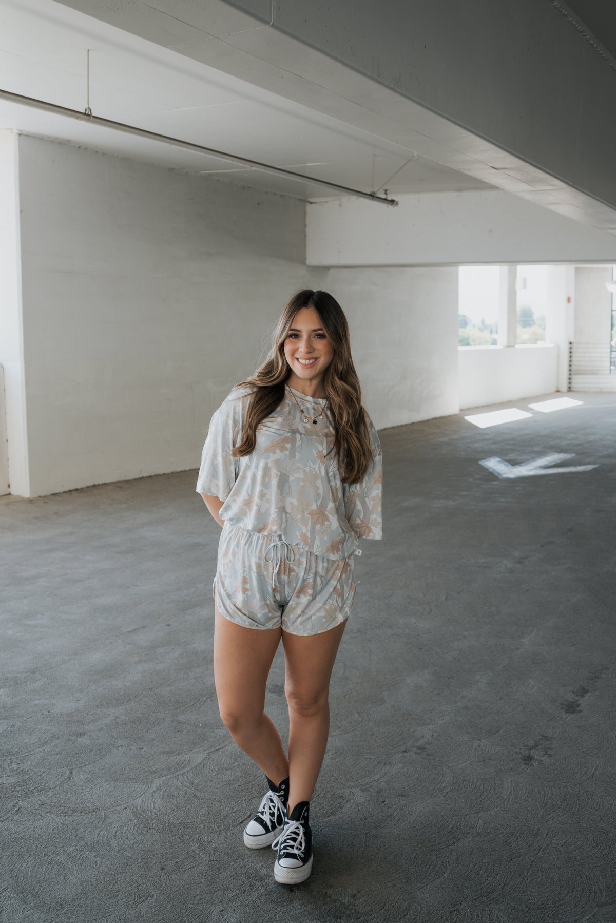 A young woman with long brown hair smiles while standing in a bright, empty parking garage, wearing the forever french baby Women’s Bamboo Short Set | Woodland and black sneakers.