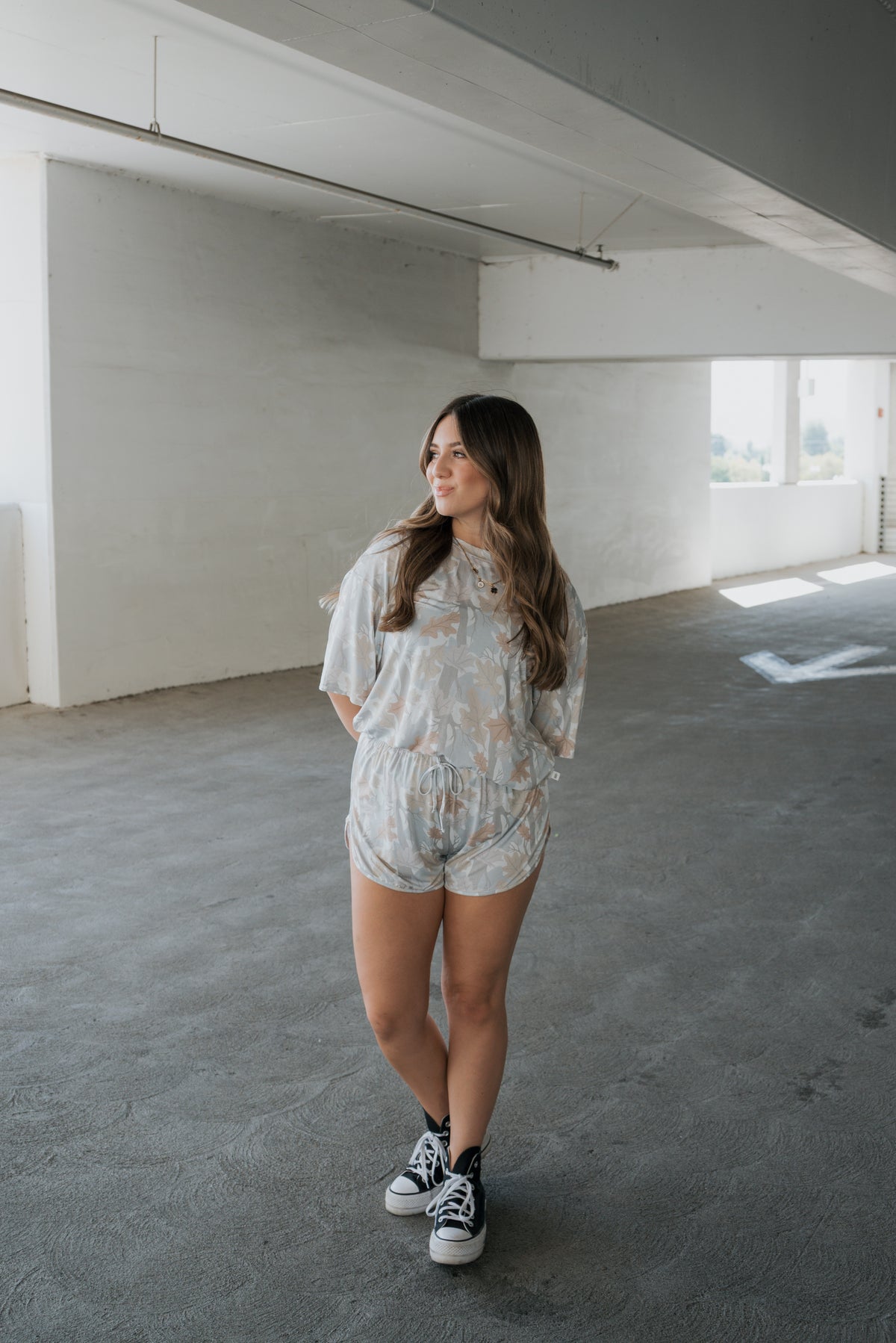 A woman stands in a bright, empty indoor space, wearing the forever french baby Women’s Bamboo Short Set | Woodland—a light-colored floral ensemble crafted from hypo-allergenic bamboo fabric. She pairs the outfit with black sneakers, her hair down, hands loosely clasped as she gazes thoughtfully to the side.