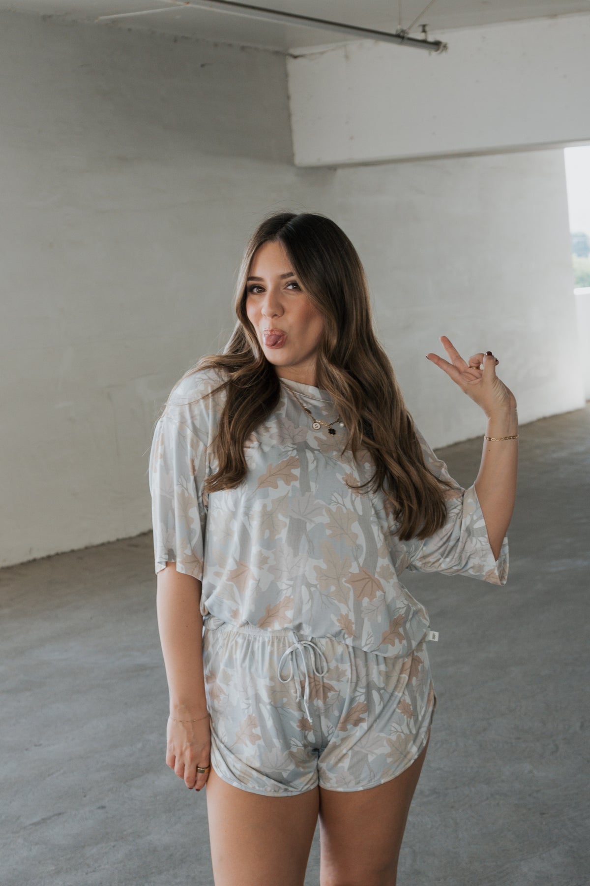 A woman in the Women’s Bamboo Short Set | Woodland by forever french baby poses indoors with long wavy hair, making a peace sign and playfully sticking out her tongue. The hypo-allergenic bamboo loungewear features a floral design and is set against a neutral background.