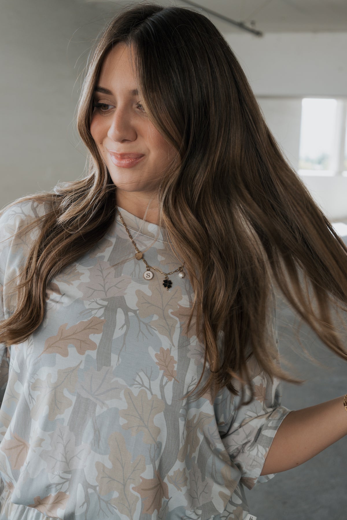 A woman with long brown hair wears the Women’s Bamboo Short Set in Woodland by forever french baby, styled with layered necklaces and smiling as she holds her hair in a softly lit room.