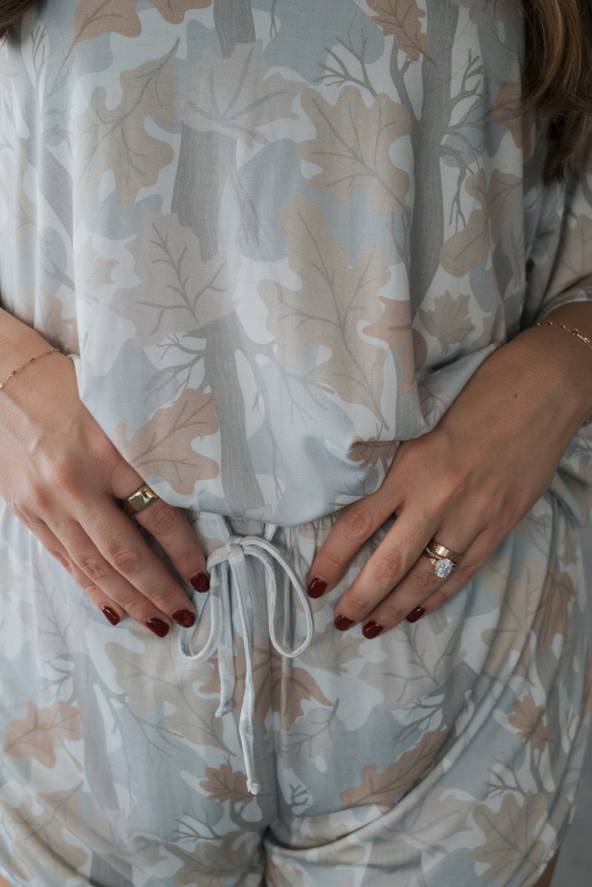 A woman wearing the forever french baby Women’s Bamboo Short Set in Woodland—a light, leaf-patterned outfit—ties the drawstring at her waist, with hands adorned in rings and red nail polish.