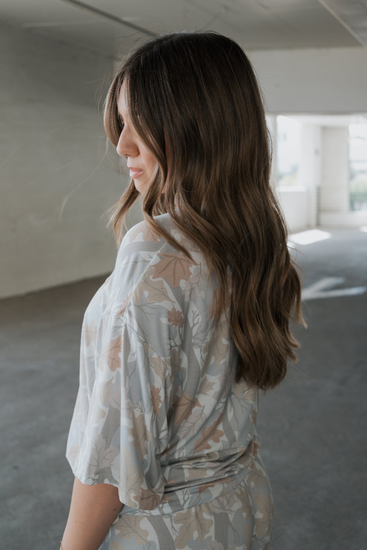 A woman with long, wavy brown hair, wearing the forever french baby Women’s Bamboo Short Set in the Woodland print, stands indoors facing sideways in soft natural light.