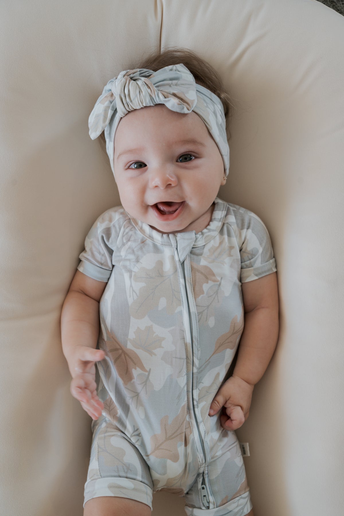 A smiling baby wearing the forever french baby Shortie Bamboo Zip in Woodland print and a matching headband lies on a soft cream cushion, looking up with one hand raised.
