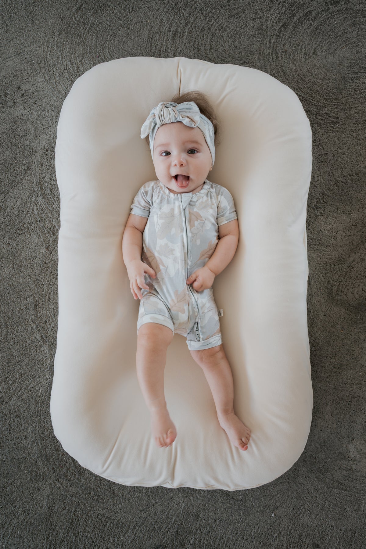 Wearing the forever french baby Shortie Bamboo Zip in Woodland, a baby with a headband lies smiling with tongue out on a cream-colored cushion atop a textured gray surface.