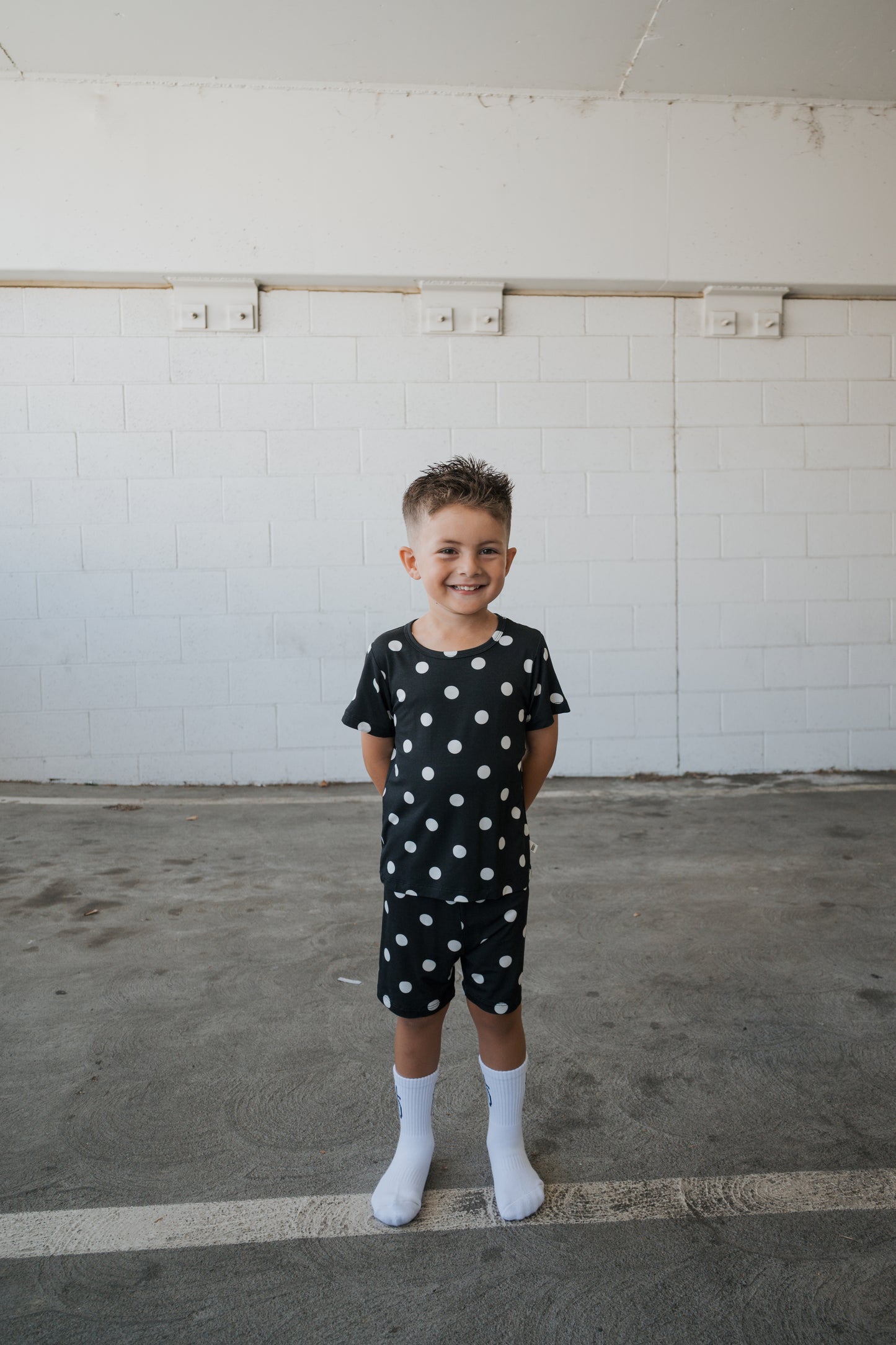 A smiling young boy stands with his hands behind his back in a parking garage, wearing the forever french baby Bamboo Short Set in B&W Polka Dot—a breathable black shirt and shorts adorned with white polka dots.
