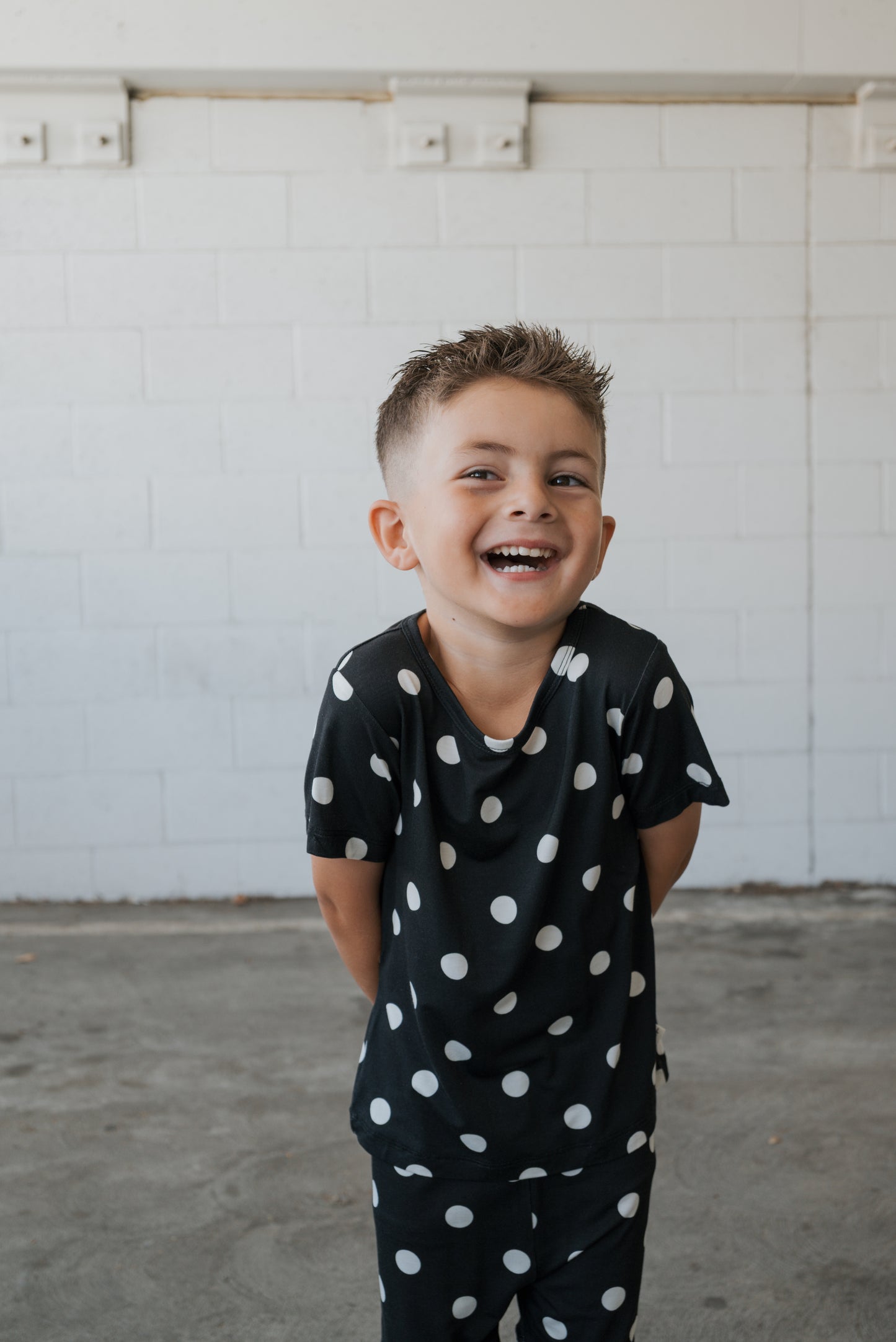 A smiling young boy with short hair stands in front of a white brick wall, hands behind his back, wearing the forever french baby Bamboo Short Set | B&W Polka Dot.