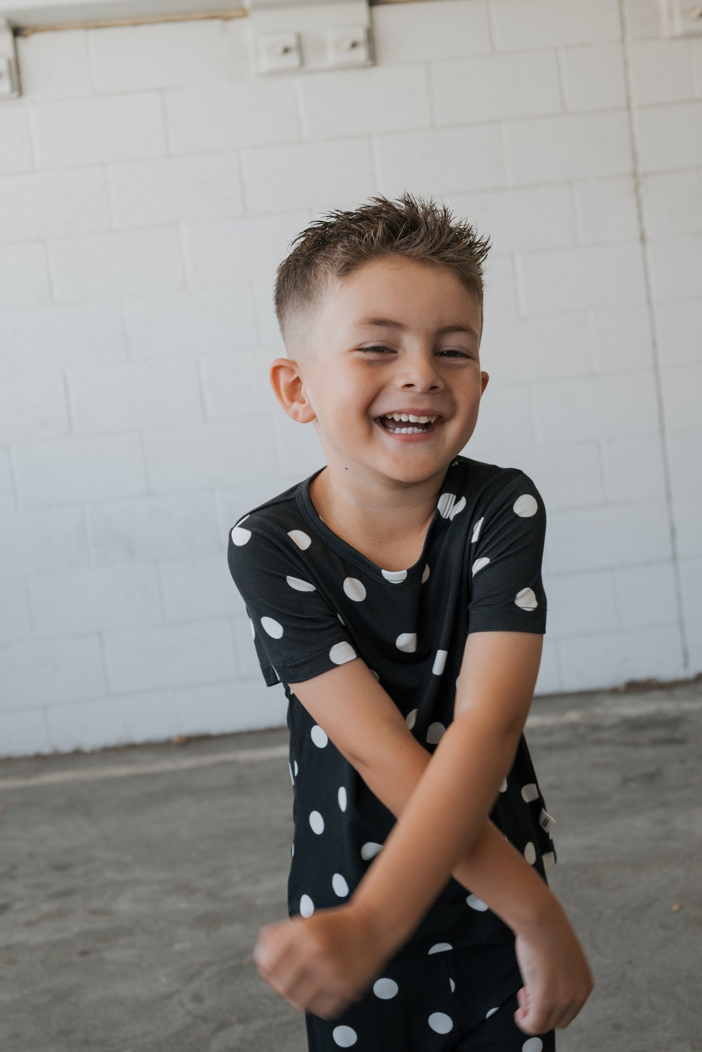 A young child with short hair, wearing the forever french baby Bamboo Short Set in the B&W Polka Dot pattern—crafted from hypo-allergenic fabric—smiles and crosses their arms while standing in front of a white brick wall.