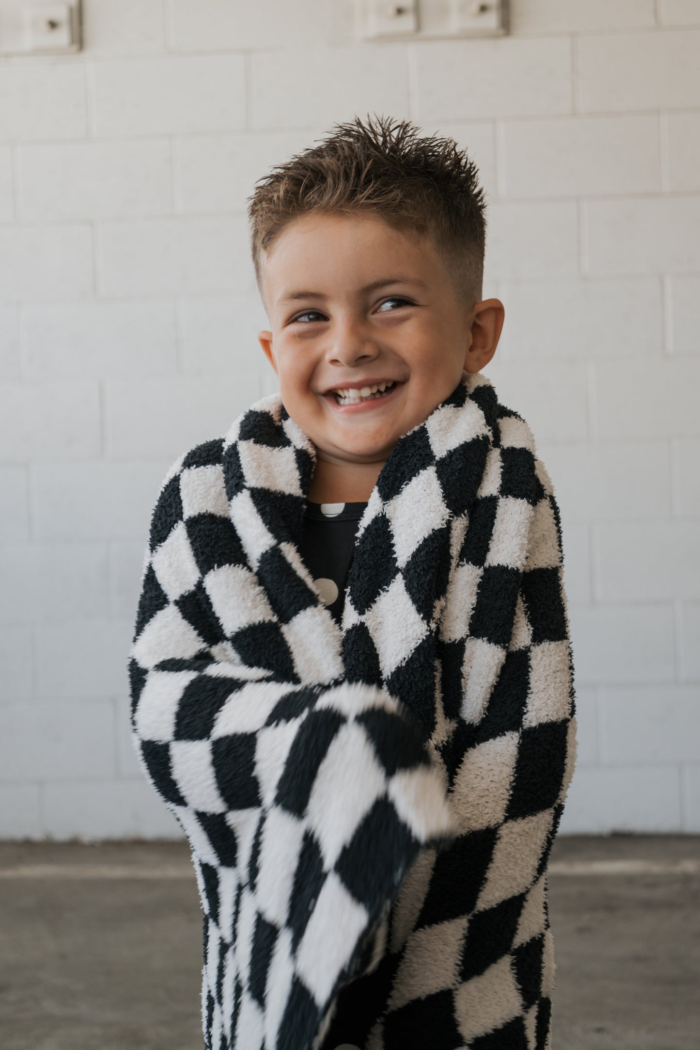 A smiling young boy stands in front of a white brick wall, wrapped in the forever french baby Plush Blanket | B&W Micro Check.
