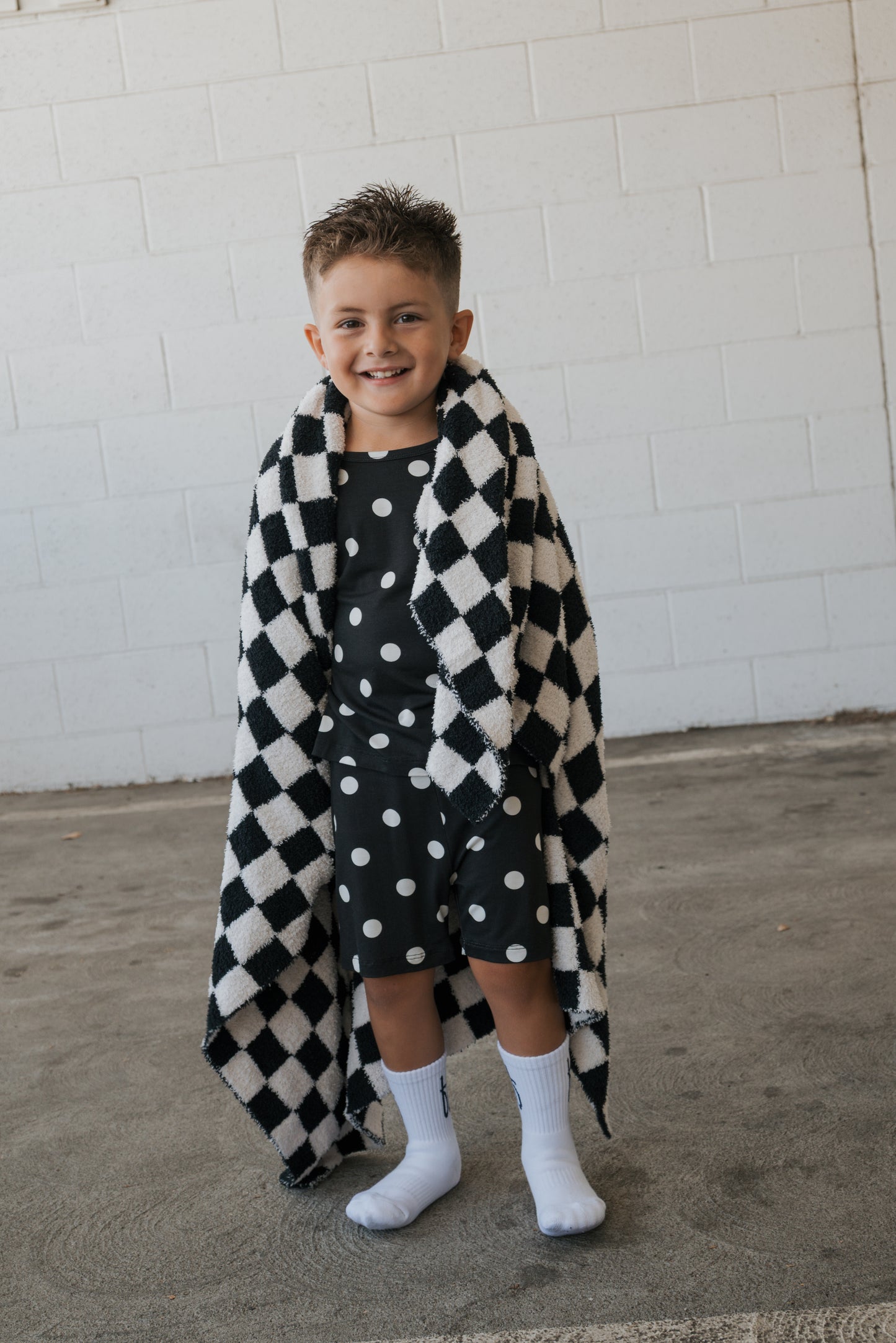 A smiling young boy stands on a concrete floor against a white brick wall, wearing the forever french baby Bamboo Short Set | B&W Polka Dot—breathable kids clothing featuring a black shirt and shorts with white polka dots—paired with white socks and wrapped in a black and white checkered blanket.