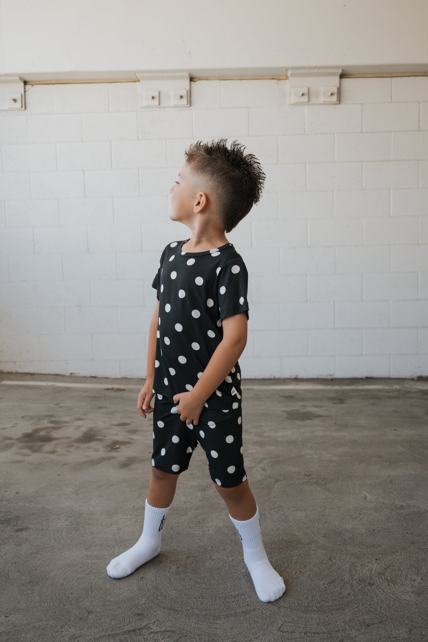 A young boy with a mohawk hairstyle wears the forever french baby Bamboo Short Set in B&W Polka Dot—hypo-allergenic clothing featuring a black outfit with white polka dots and white socks—as he stands on concrete and looks to his left against a white brick wall.