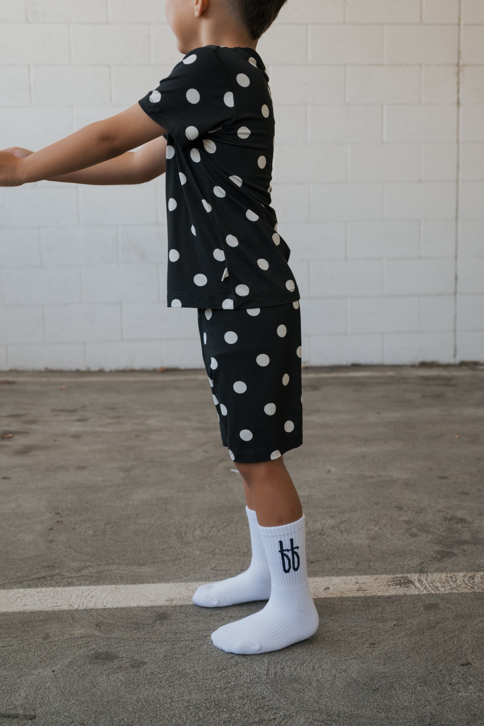 A child stands sideways against a white brick wall and concrete floor, wearing the forever french baby Bamboo Short Set | B&W Polka Dot—a stylish yet comfortable set of hypo-allergenic bamboo pajamas featuring a black shirt and polka-dot shorts—and white socks adorned with a dark “bb” logo.