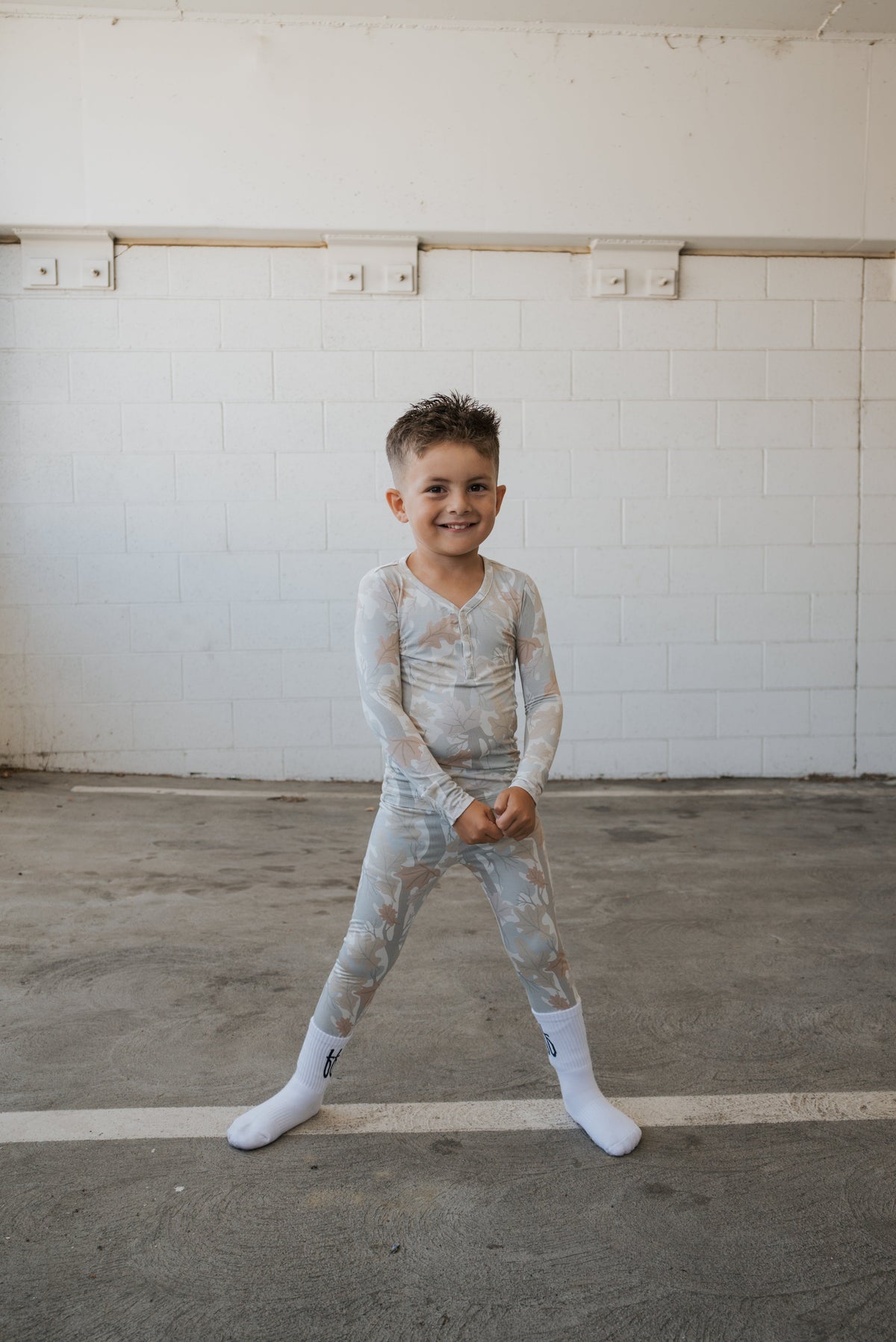 A young boy with short hair stands smiling in forever french baby’s Bamboo Two Piece Pajamas | Woodland, soft and hypoallergenic kids sleepwear, paired with white socks and featuring a subtle woodland pattern. He is posed indoors against a white brick wall and concrete floor.