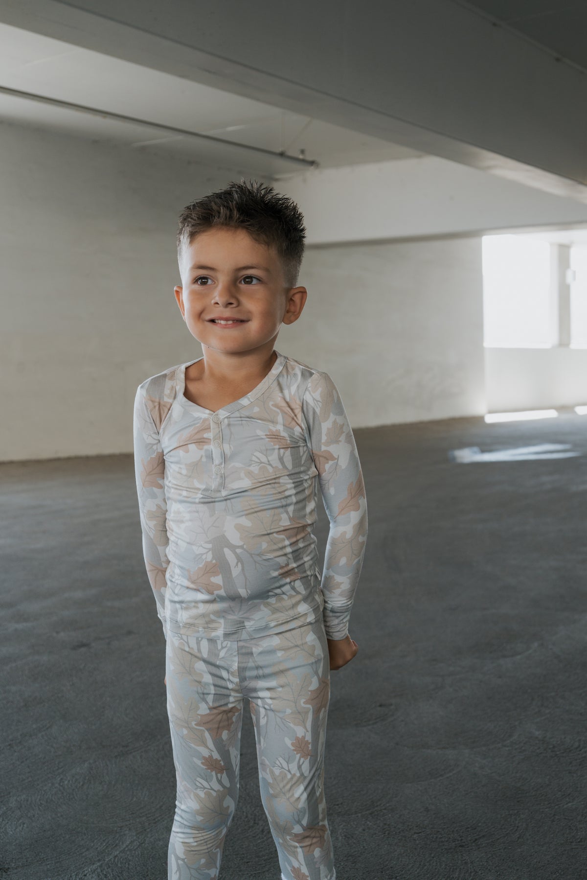 A young child with short hair smiles while wearing the forever french baby Bamboo Two Piece Pajamas in the Woodland print, standing in a large, empty, well-lit room.