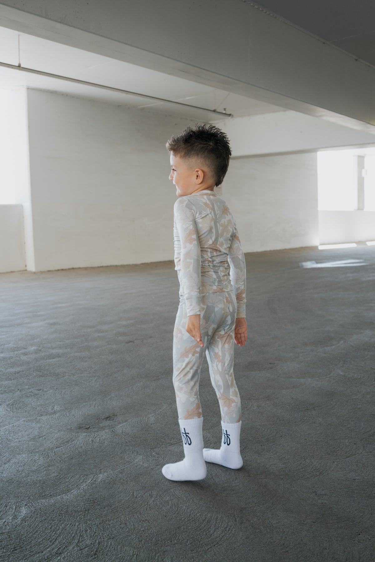 A young child wearing forever french baby’s Bamboo Two Piece Pajamas in the Woodland pattern and white socks stands on a gray floor in a bright, empty room, looking to the side.