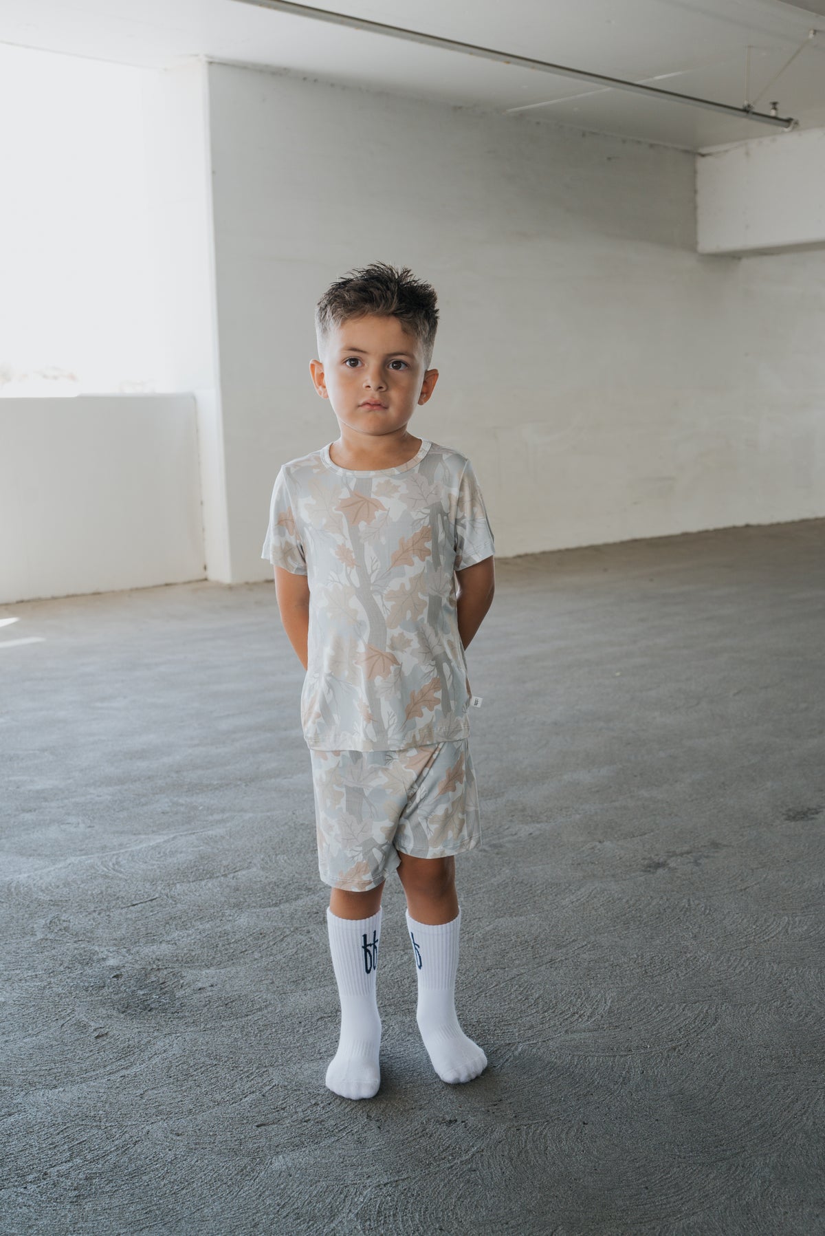 A young boy with short brown hair stands indoors on a gray floor, wearing the forever french baby Bamboo Short Set | Woodland—a matching light-colored shirt and shorts set paired with white knee-high socks. He looks slightly serious, with his hands behind his back, embodying the simple comfort of hypoallergenic kids sleepwear.