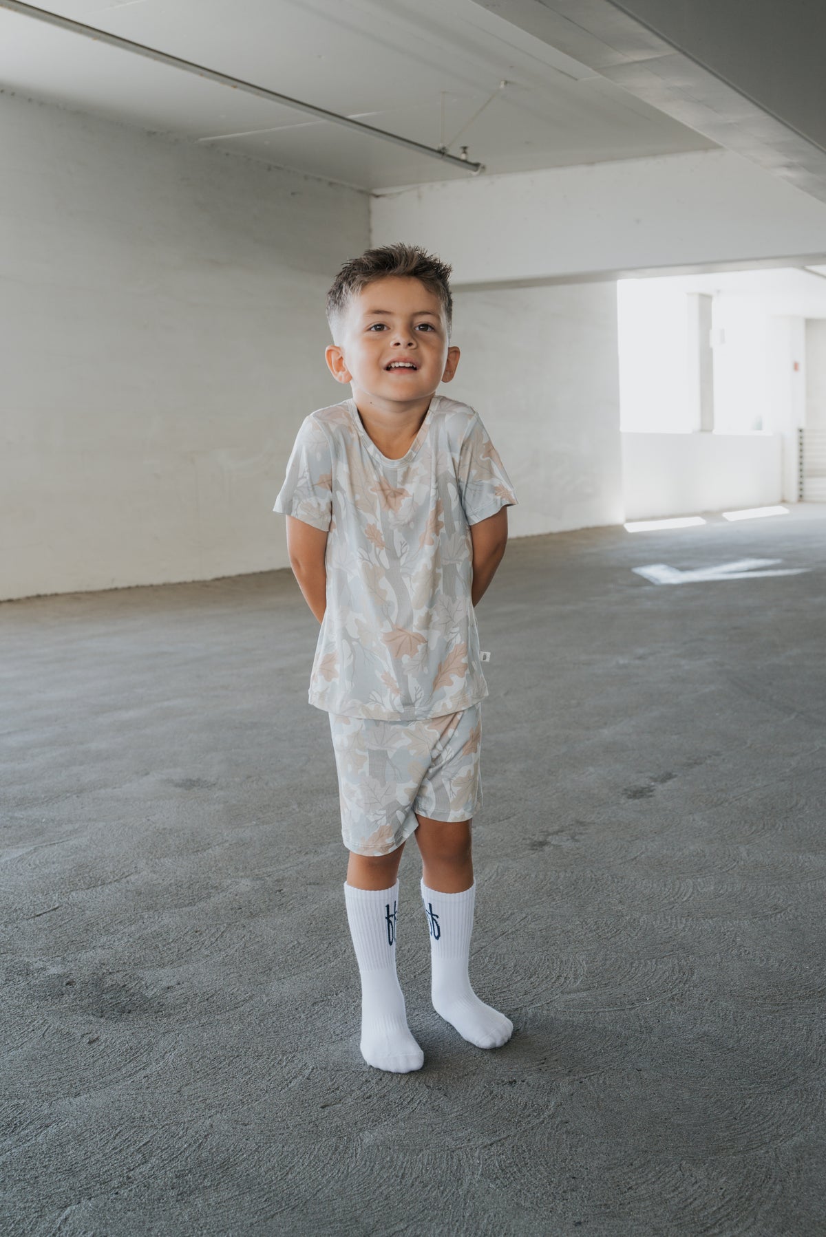 A young boy stands indoors on a gray floor, smiling with his hands behind his back. He is wearing the forever french baby Bamboo Short Set in the Woodland print—matching, light-colored pajamas crafted from hypoallergenic bamboo fabric that keeps him comfortable all day long.