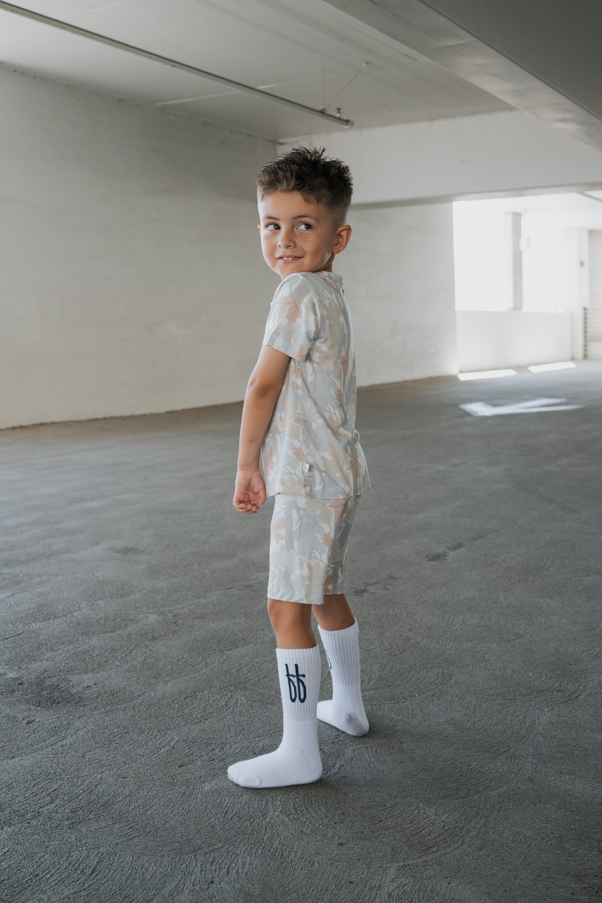 A young boy in the forever french baby Bamboo Short Set | Woodland stands on a concrete floor in a bright, empty room, looking over his shoulder and smiling—perfectly cozy in his soft, hypoallergenic kids sleepwear.
