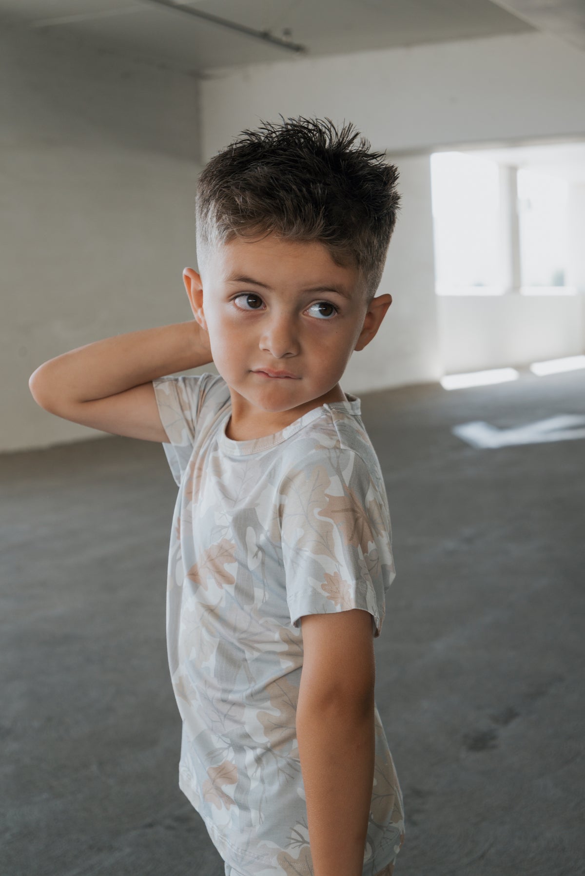 A young boy with short brown hair stands indoors, looking to the side with his hand resting on the back of his neck, wearing the Bamboo Short Set in Woodland by forever french baby.