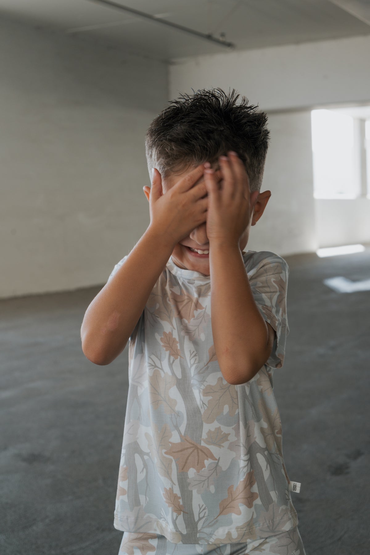 A young boy stands in a bright, empty room, smiling while covering his face with both hands as if playing hide and seek. He’s dressed in the forever french baby Bamboo Short Set | Woodland, perfect for comfortable, hypoallergenic kids sleepwear.