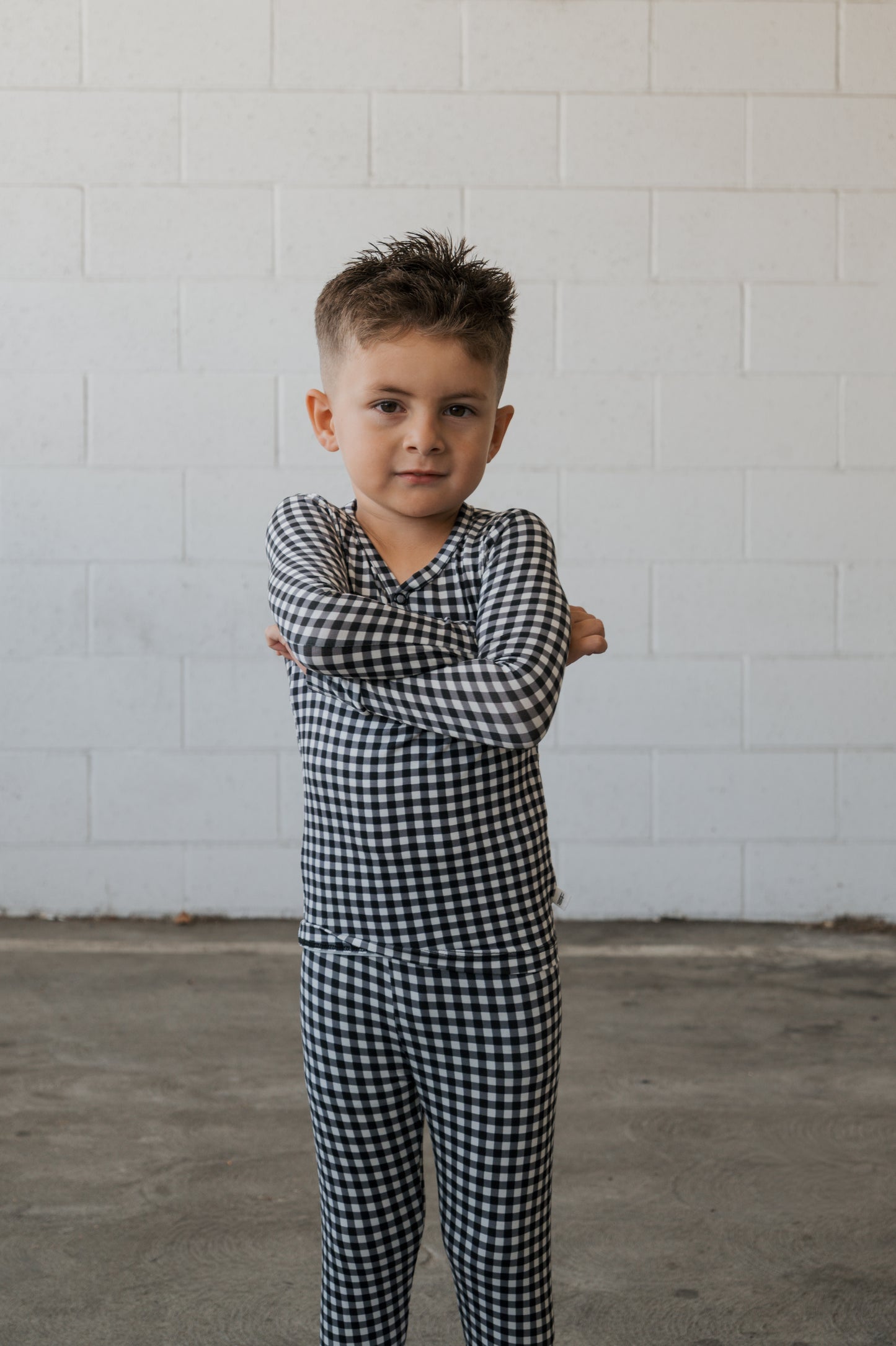 A young child with short hair stands confidently on a concrete floor, arms folded, wearing the forever french baby Bamboo Two Piece Pajamas in Black Gingham, in front of a white brick wall.