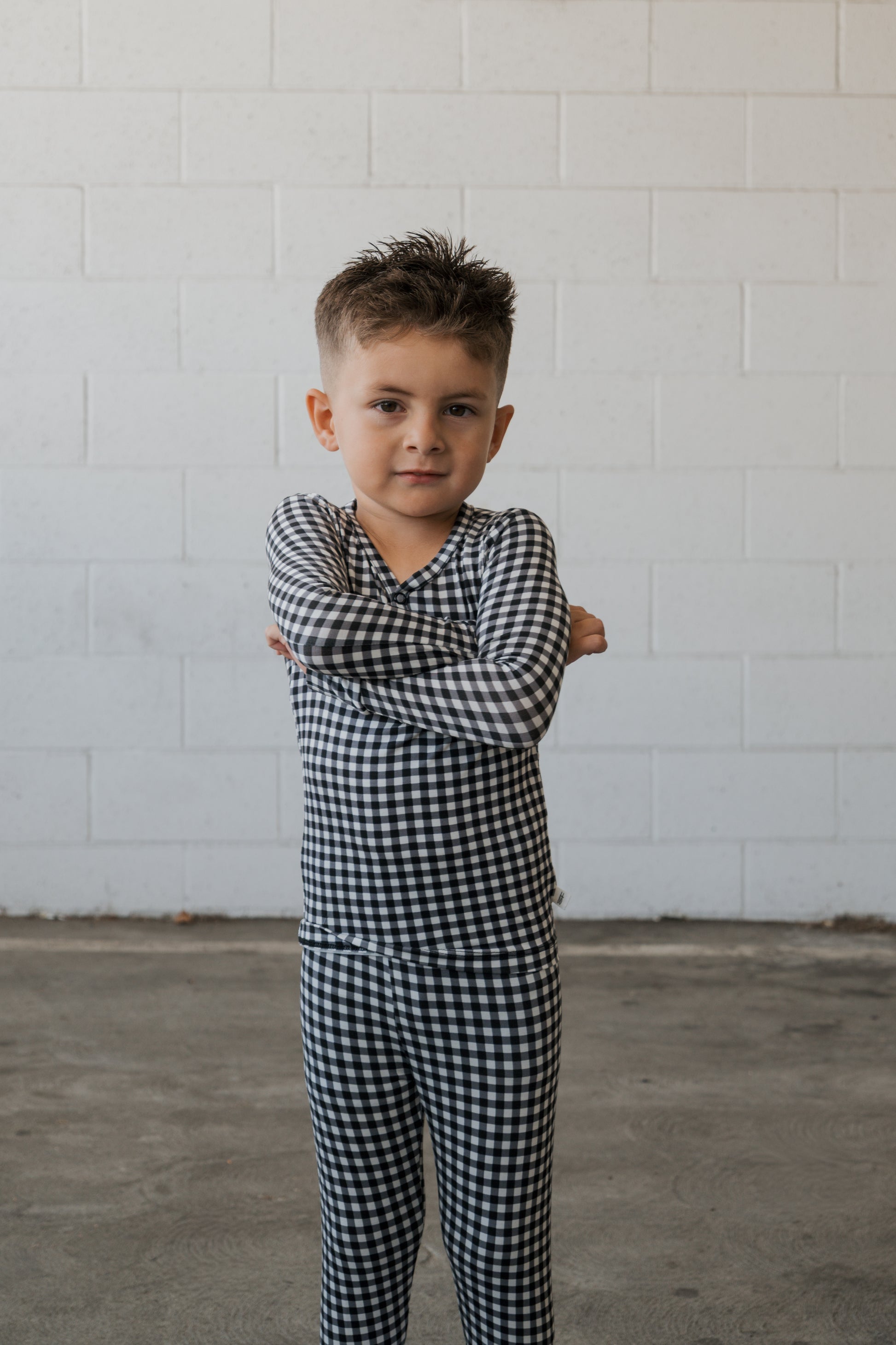 A young child with short hair stands confidently on a concrete floor, arms folded, wearing the forever french baby Bamboo Two Piece Pajamas in Black Gingham, in front of a white brick wall.