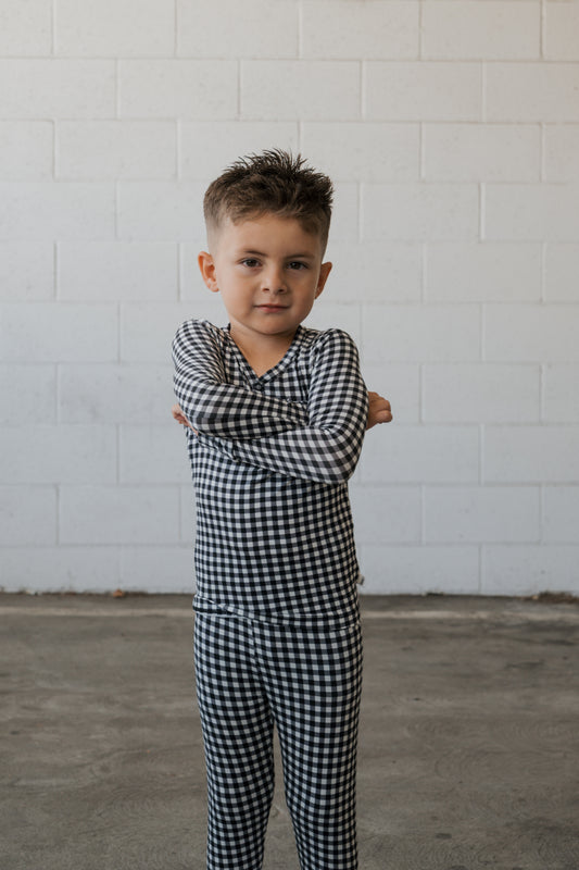 A young child with short hair stands confidently on a concrete floor, arms folded, wearing the forever french baby Bamboo Two Piece Pajamas in Black Gingham, in front of a white brick wall.