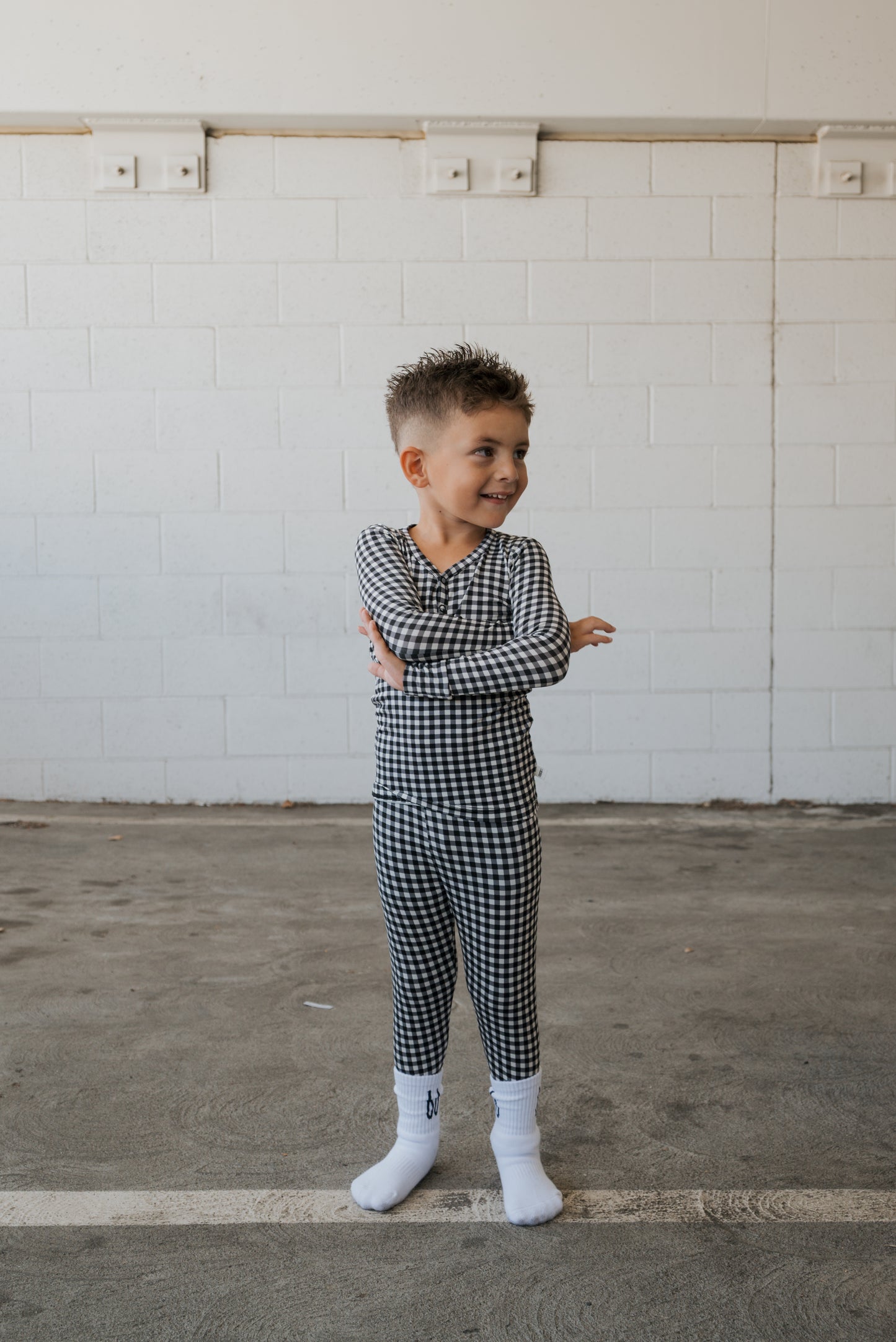 A young child wearing forever french baby’s Bamboo Two Piece Pajamas in Black Gingham and white socks stands on a concrete floor, smiling and crossing their arms, with a white brick wall in the background.