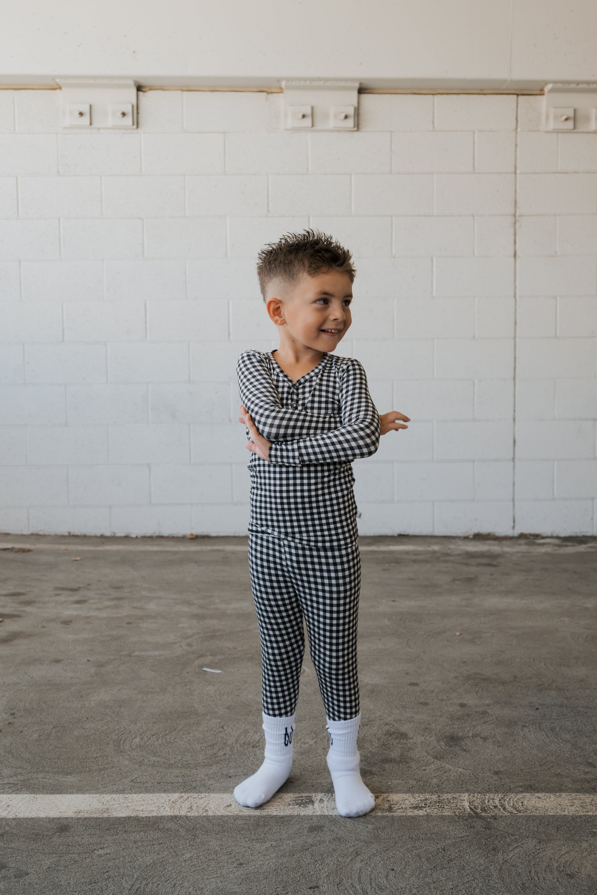 A young child wearing forever french baby’s Bamboo Two Piece Pajamas in Black Gingham and white socks stands on a concrete floor, smiling and crossing their arms, with a white brick wall in the background.