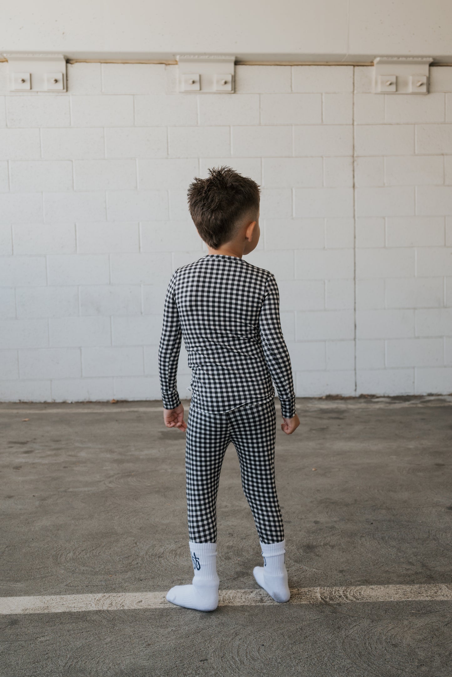 A young boy wearing forever french baby’s Bamboo Two Piece Pajamas in Black Gingham and white socks stands indoors on a concrete floor, facing a white brick wall with his back to the camera.