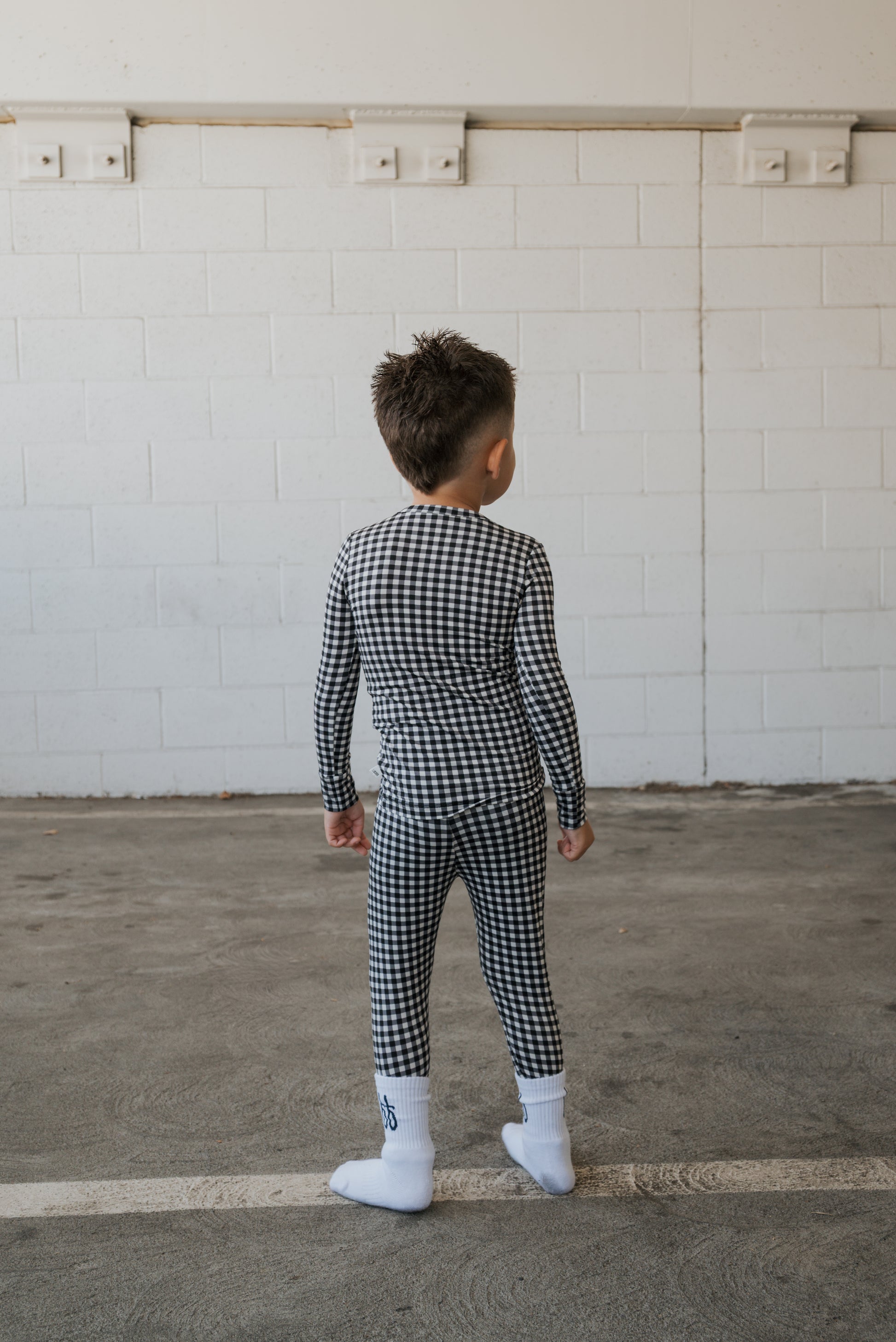 A young boy wearing forever french baby’s Bamboo Two Piece Pajamas in Black Gingham and white socks stands indoors on a concrete floor, facing a white brick wall with his back to the camera.