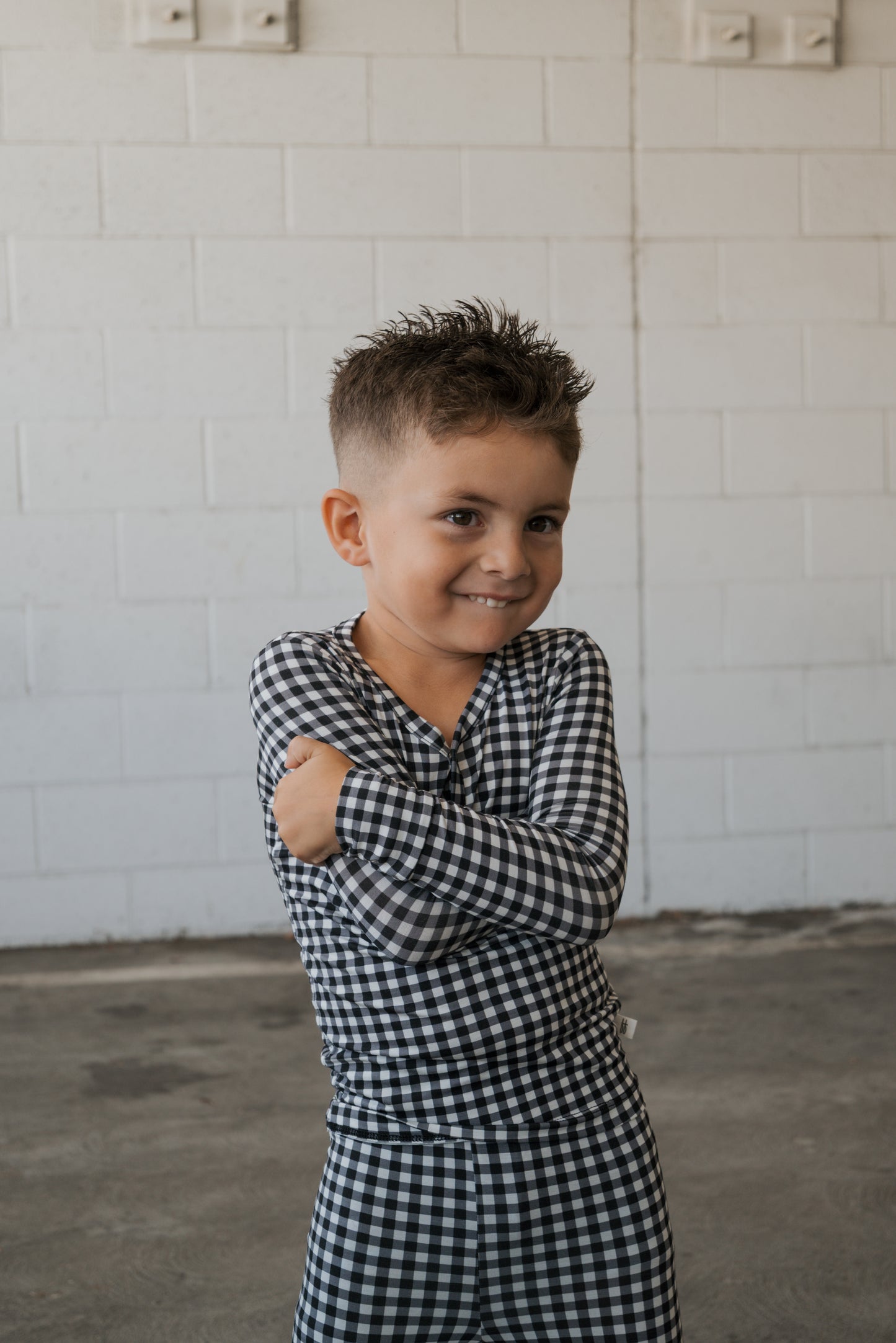 A young boy smiles with his arms crossed, wearing the forever french baby Bamboo Two Piece Pajamas in Black Gingham, standing in front of a white brick wall.
